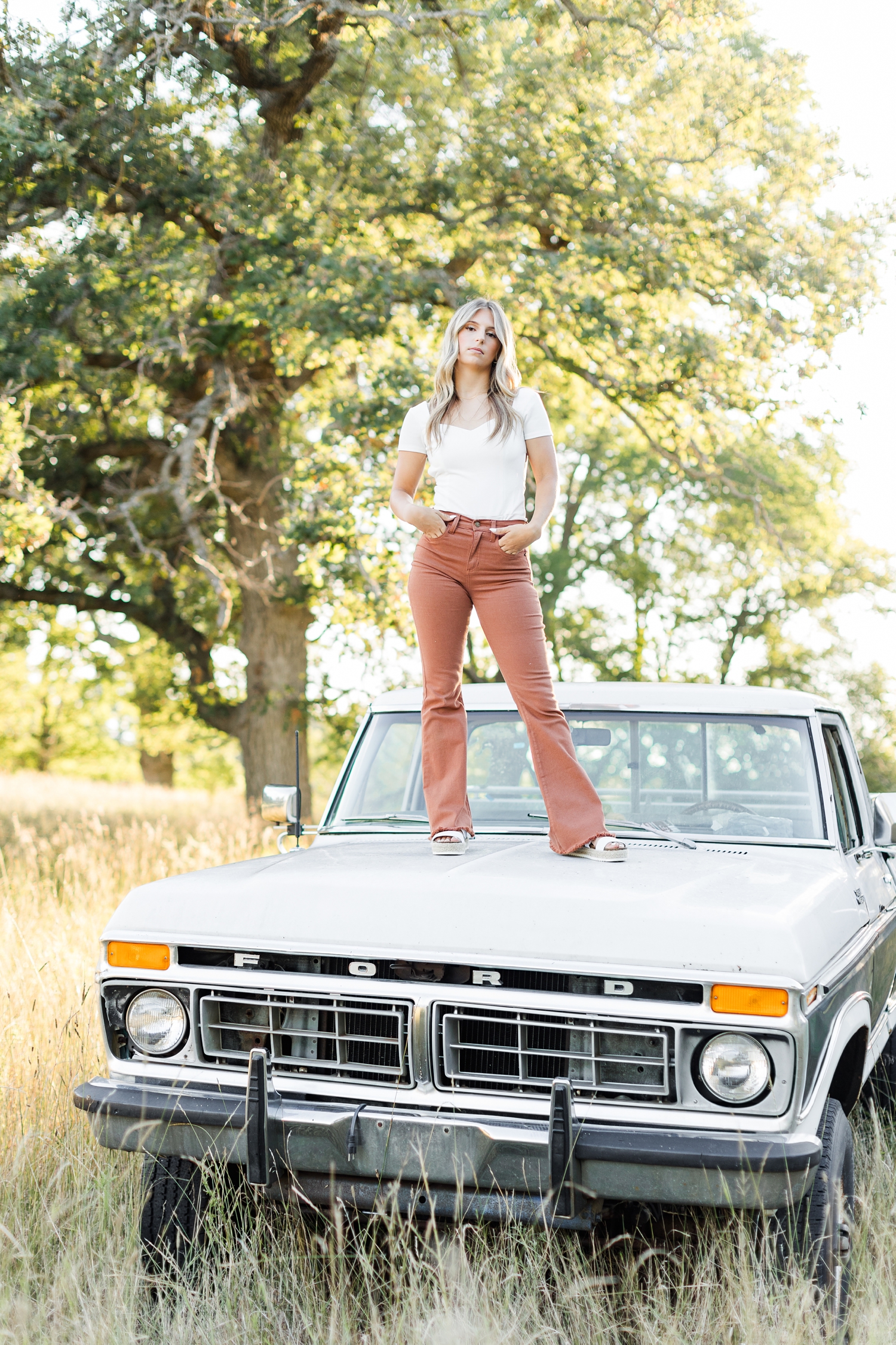 Eden, wearing burnt orange bell bottoms, stands on the hood of a 1970's Ford High Boy in the middle of a grassy field | CB Studio