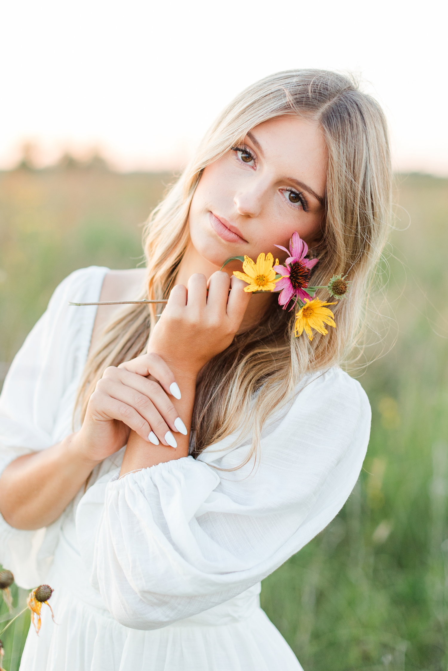 Eden stands in a wildflower field at golden hour wearing a white dress holding flowers to her cheek | CB Studio