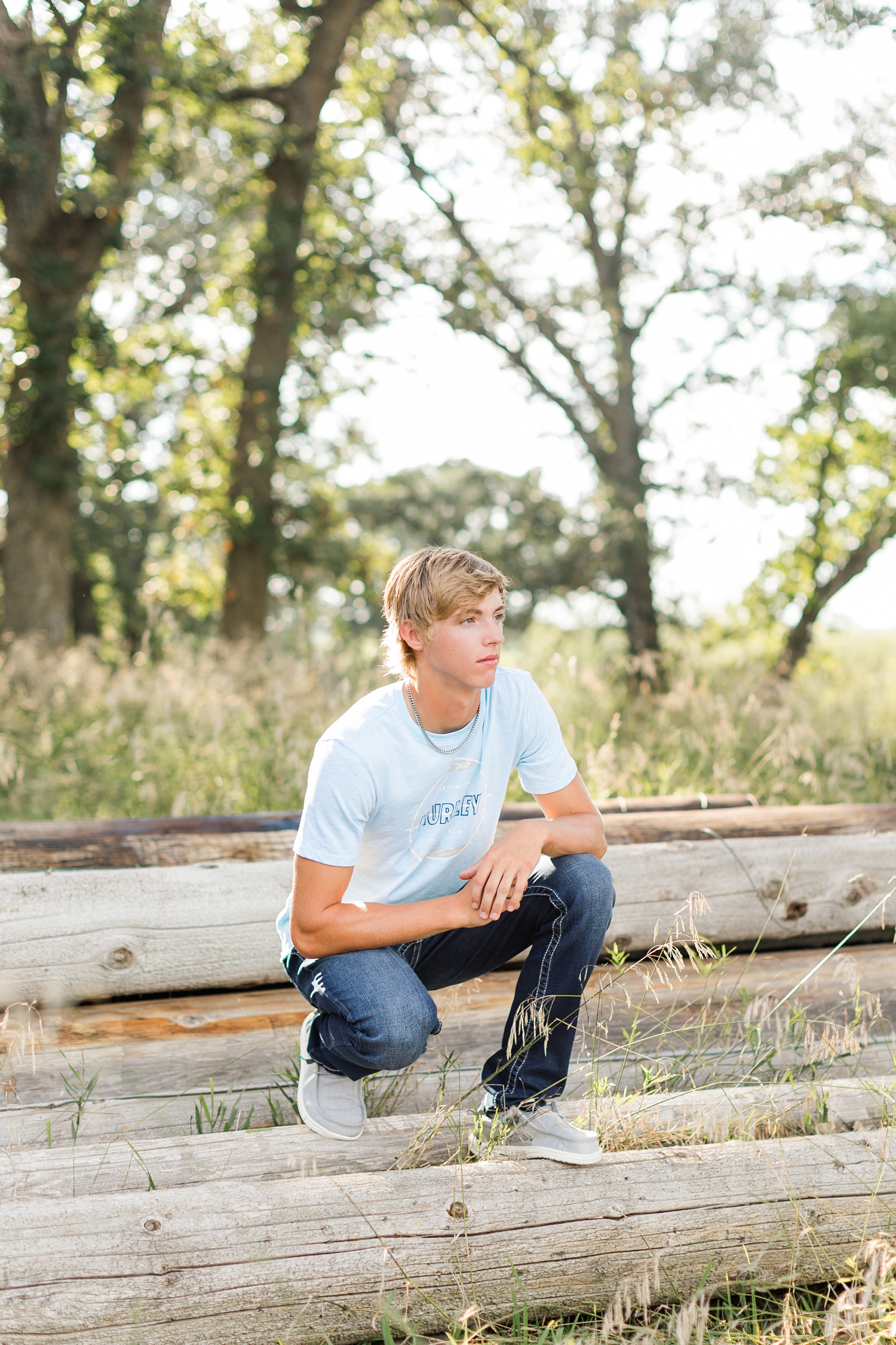 Justin squats on a stack of wood logs in the middle of a grassy field | CB Studio