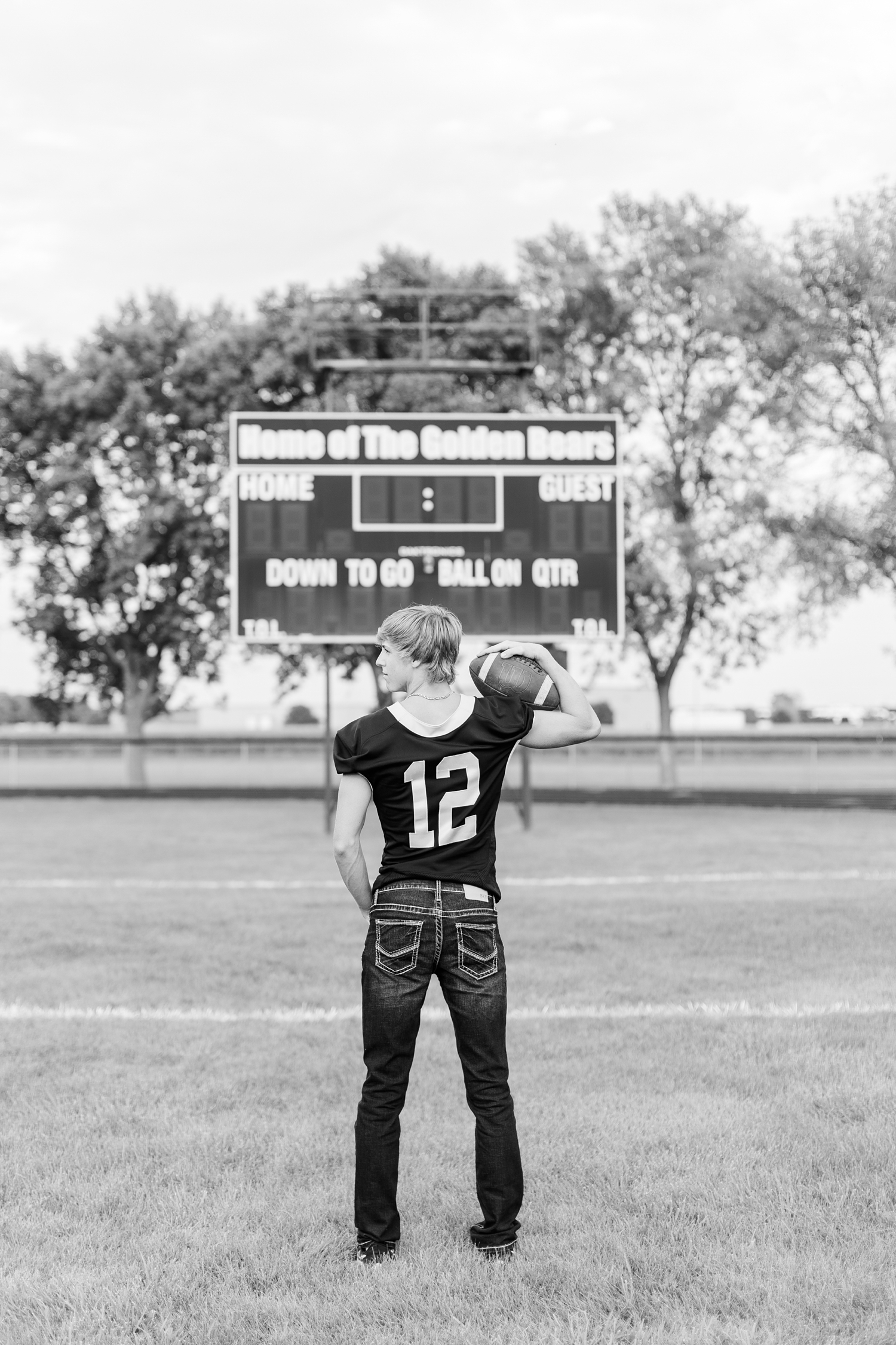 Justin stands on the football field and holds a football on his shoulder as he faces the scoreboard | CB Studio