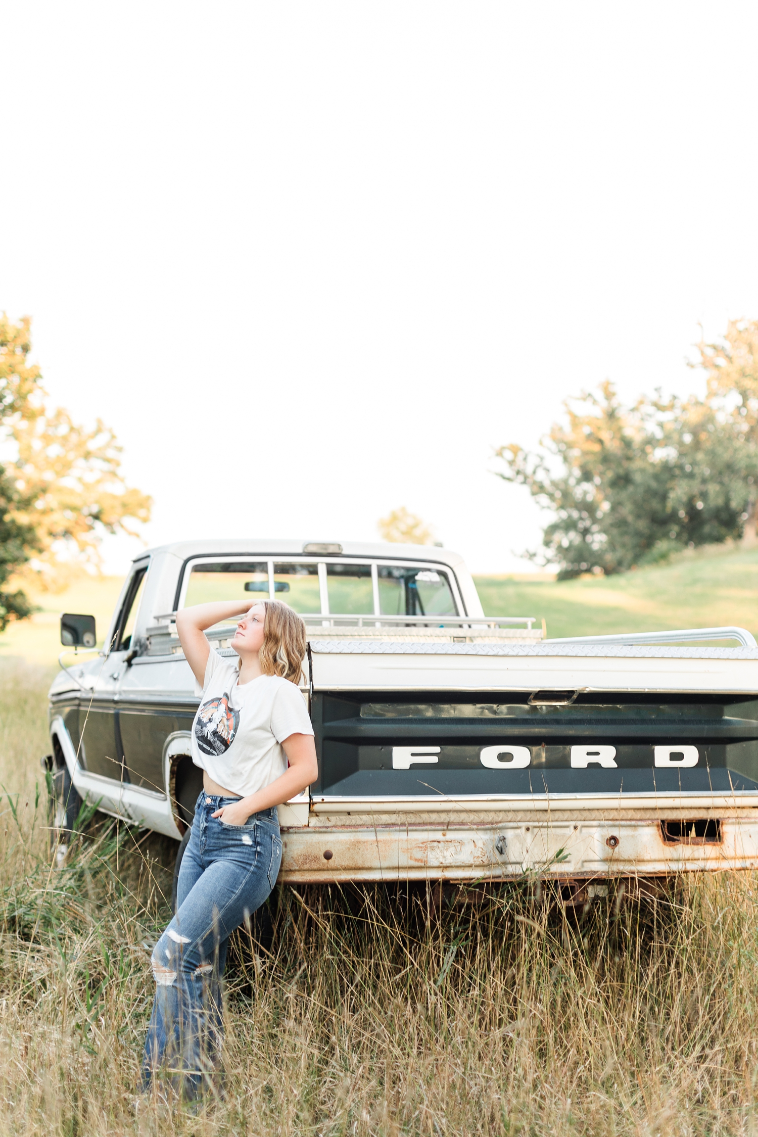 Kenzie leans on the back of a 70's Ford Highboy truck in the middle of a grassy field and looks up to the sky | CB Studio