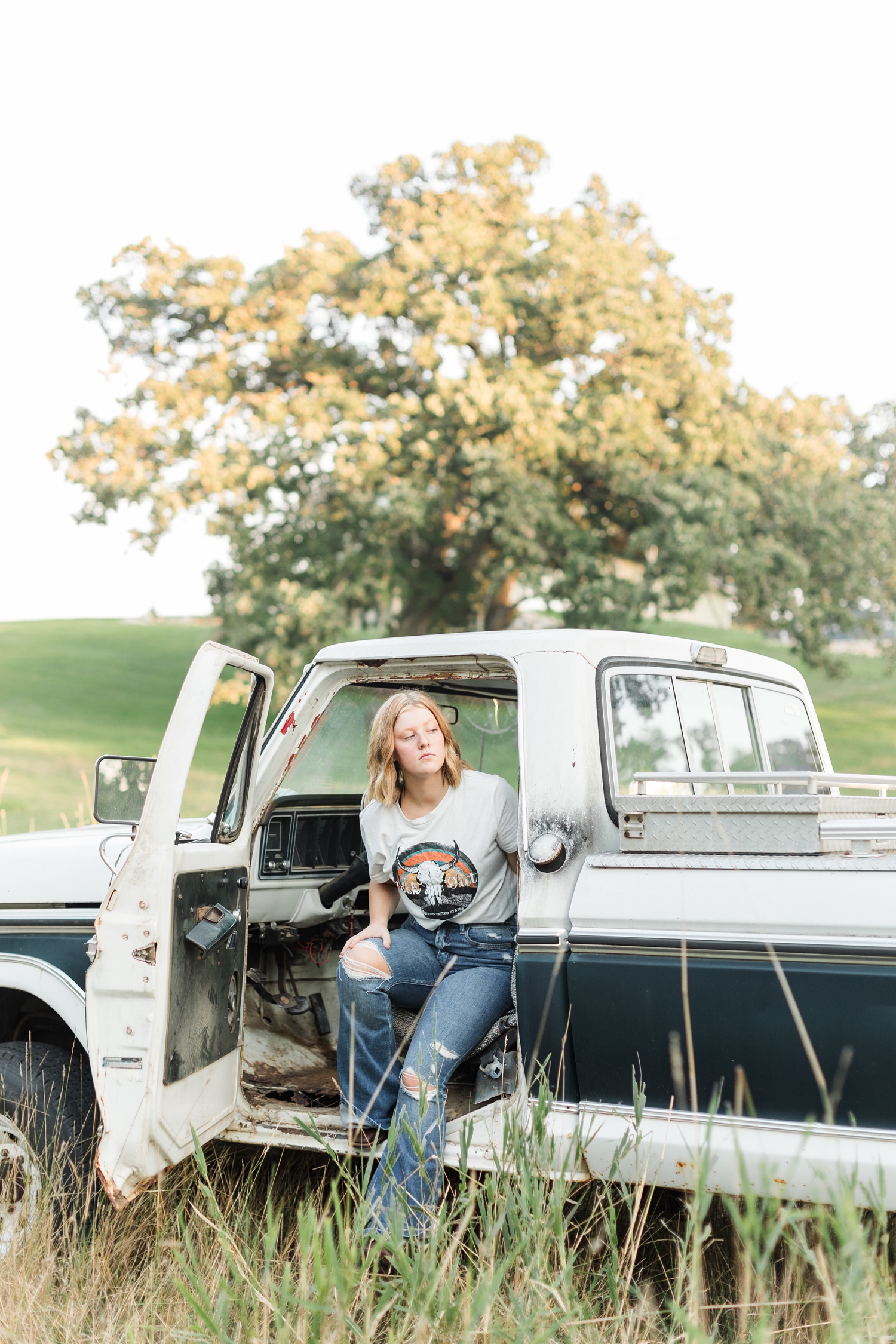 Kenzie sits in the driver's seat of a 70's Ford Highboy truck in the middle of a grassy field while looking to the back of the truck | CB Studio
