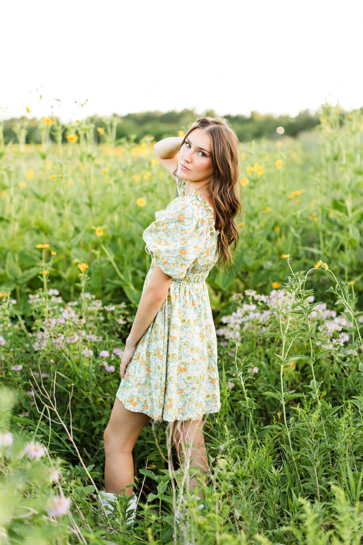 Mallory looks over her shoulder while standing in a field of yellow wildflowers wearing a green and yellow floral dress | CB Studio