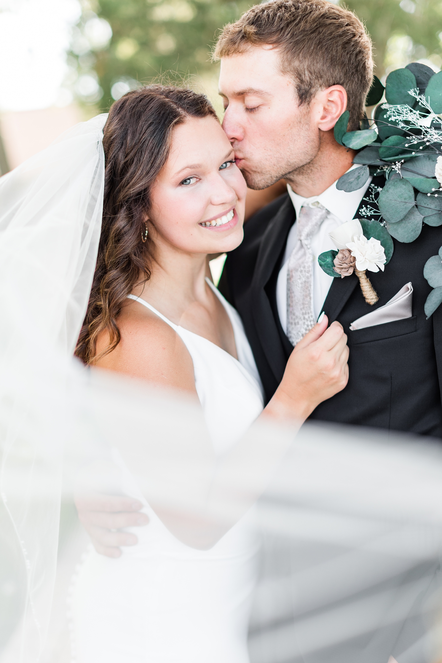 Grant kisses Alyssa on her cheek as her veil sweeps in front at 5 Island Campground in Emmetsburg, IA | CB Studio