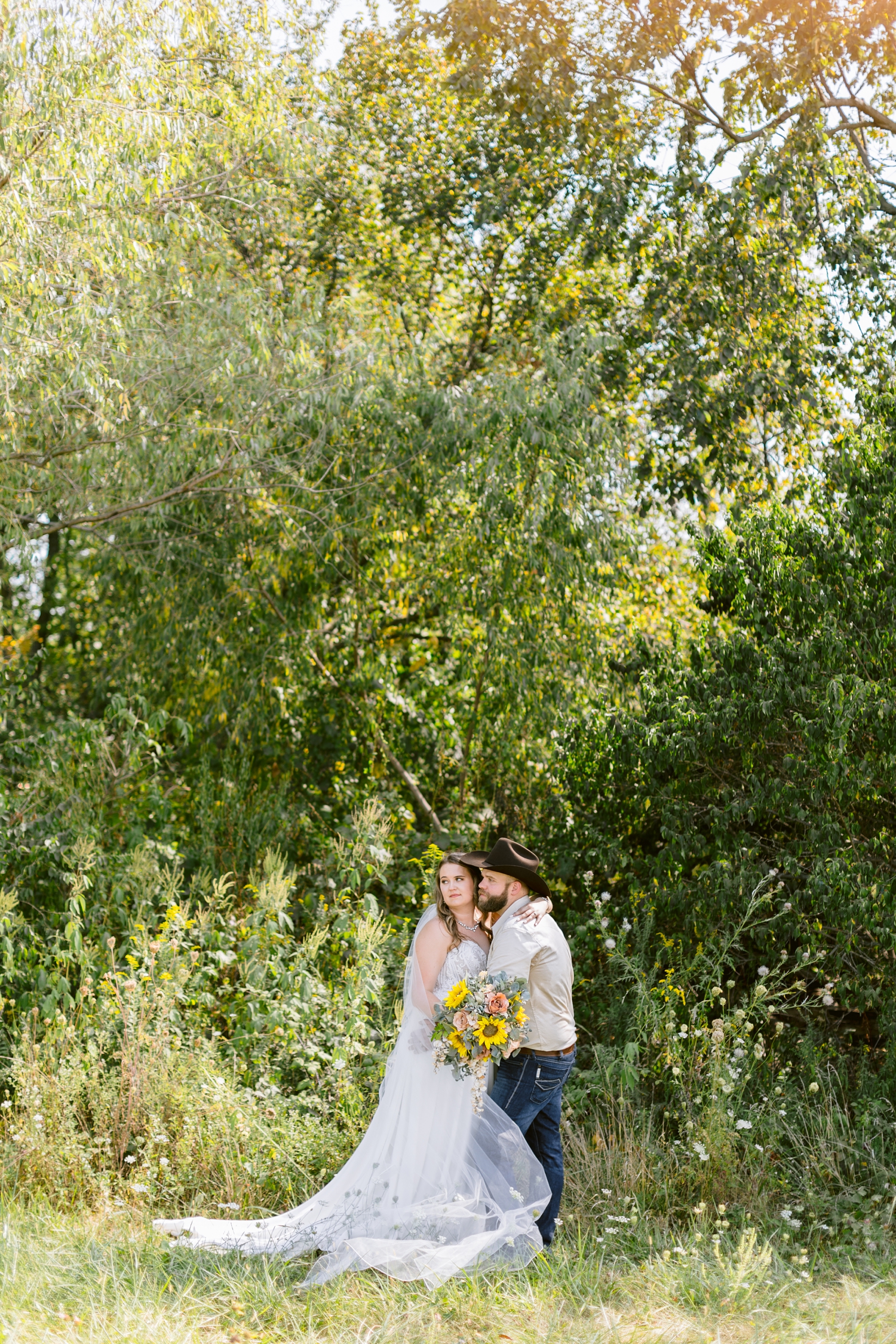Megan and Matt embrace and look off in the distance in a grassy, flower field in Knoxville, IA | A rustic, country wedding with western touches | CB Studio