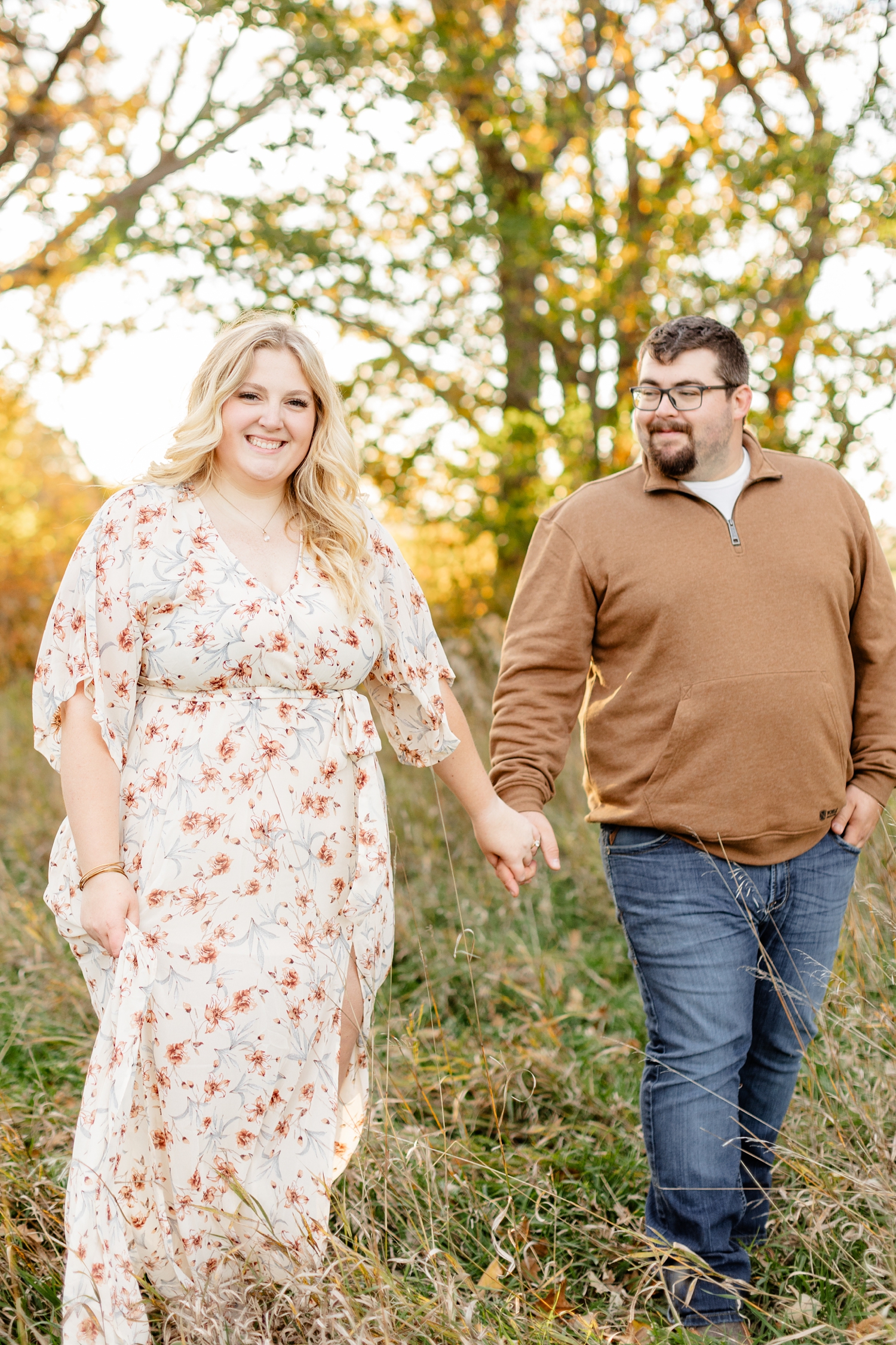 Halie and Nick walk hand in hand in an Iowa grassy pasture with peak fall oak trees in the background | CB Studio