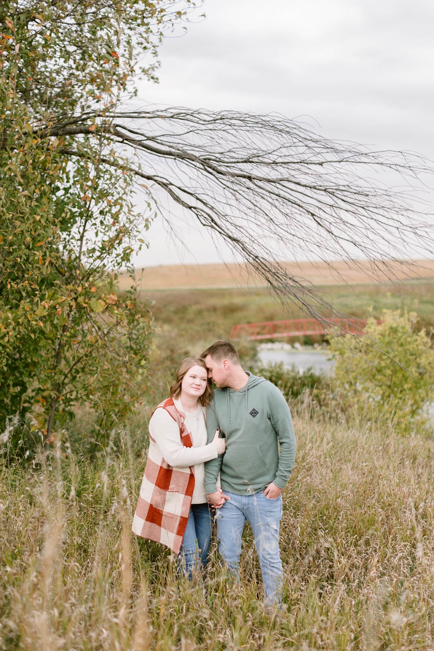 Justin nuzzles Kylie snuggle in the middle of a prairie grassy field with a red bridge in the background at Water's Edge Nature Center | CB Studio