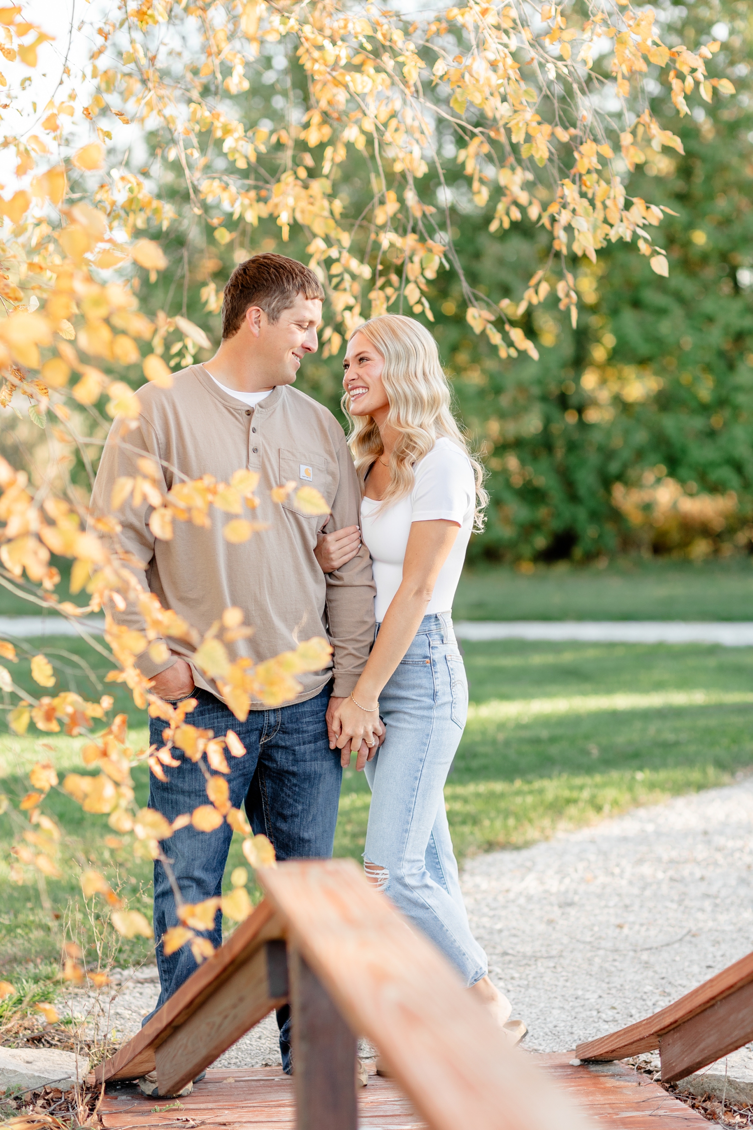Reed and Nicole hold hands and smile at each other underneath a fall colored birch tree along a bridge path at River Vally Orchards & Winery | CB Studio