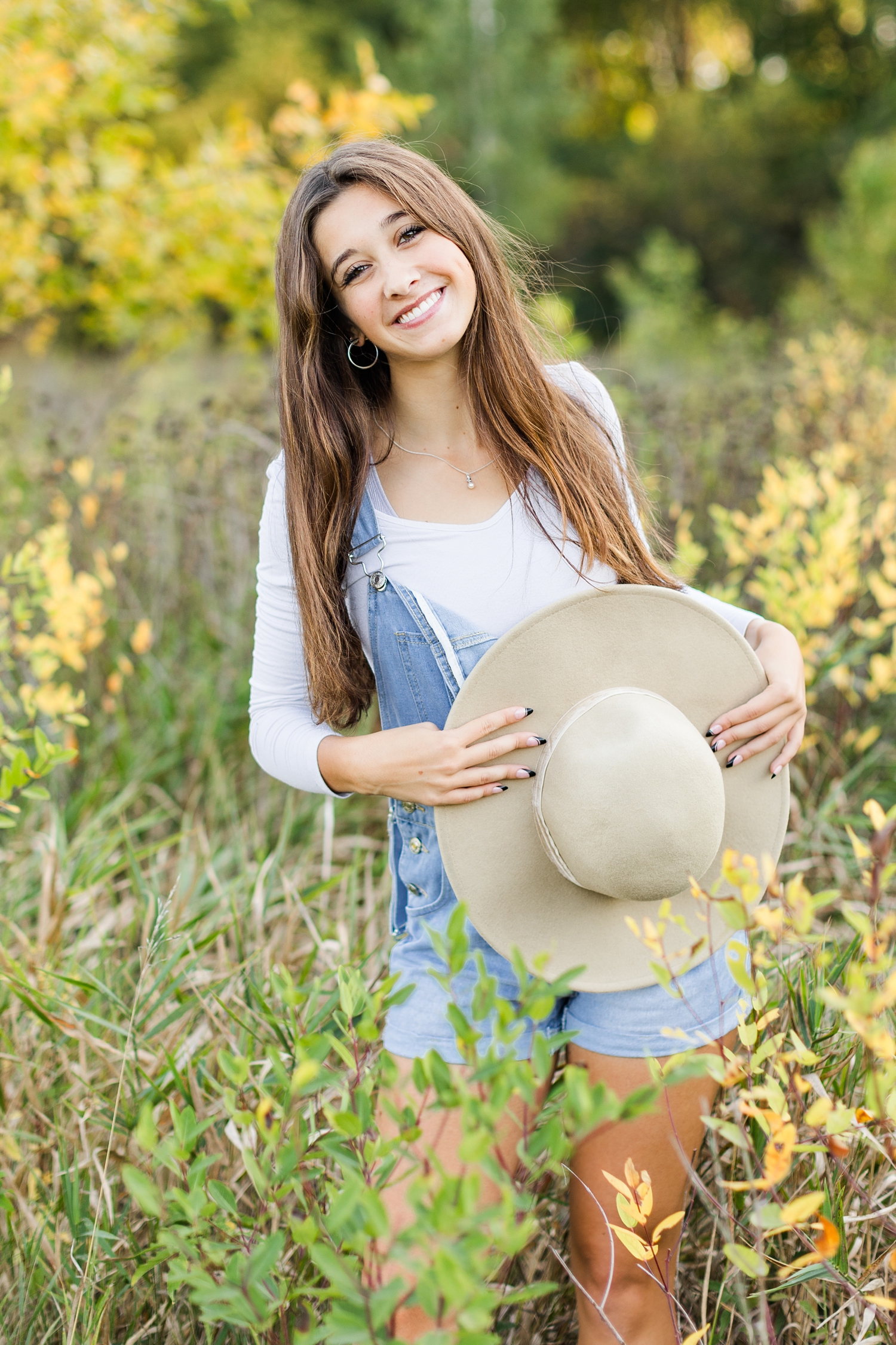 Sarah holds her wide brim hat in the middle of a grassy field surrounded by yellow fall leaves | CB Studio