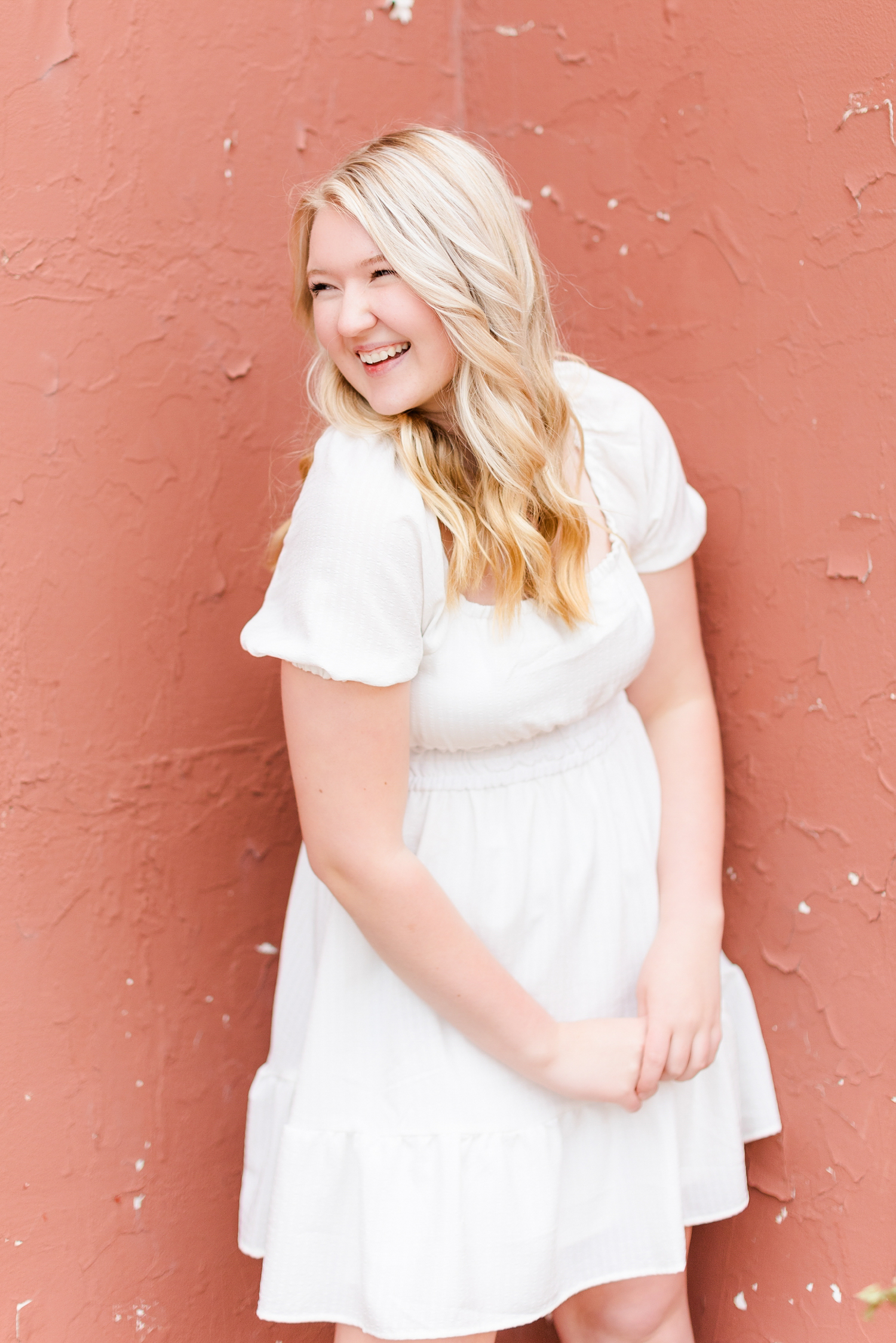 Jayden, wearing a white dress, laughs over her shoulder has she stands in front of a pink wall in downtown Algona, IA | CB Studio