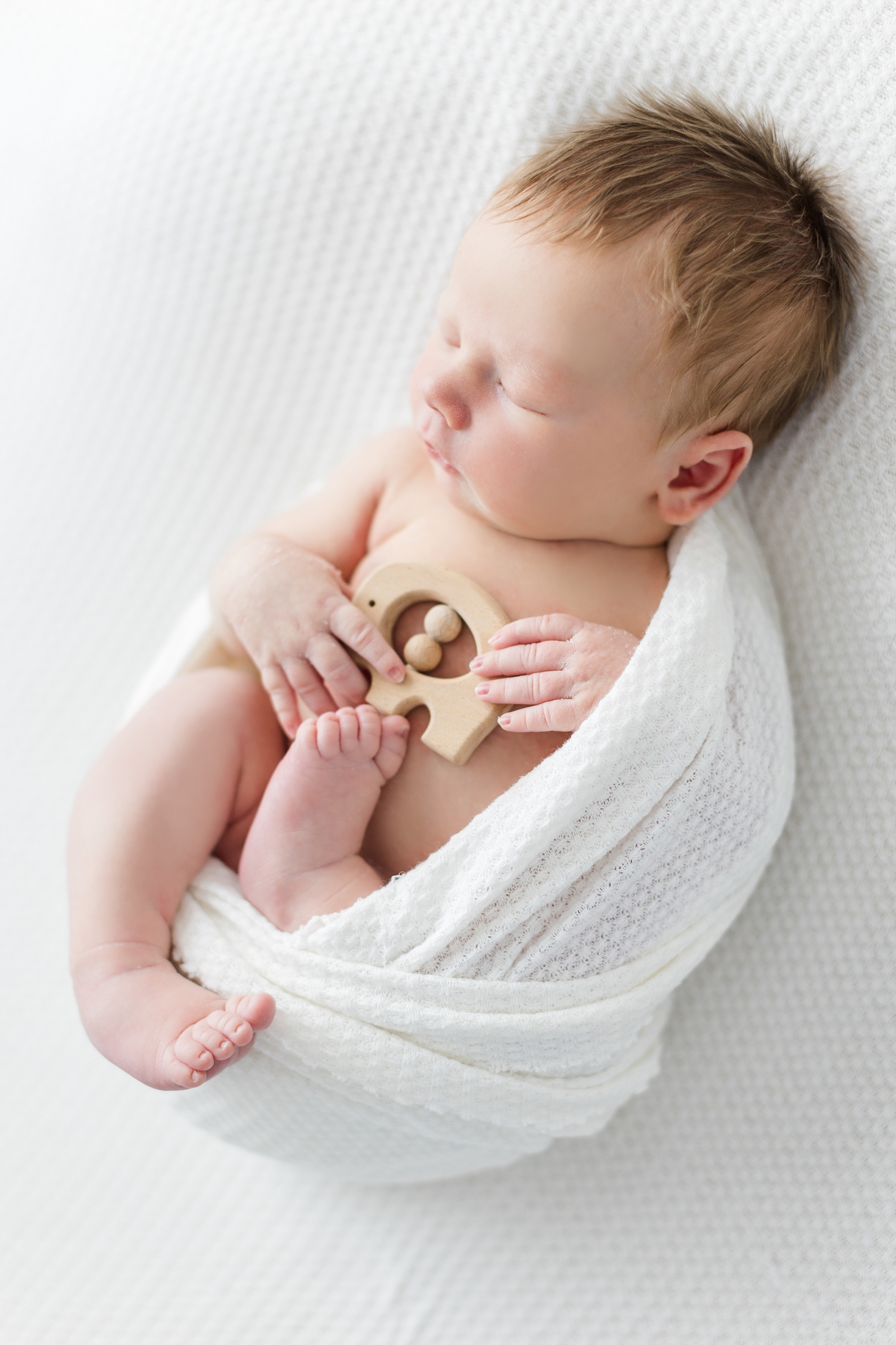 Baby Lane sleeps on an all white, waffle knit background wrapped in a matching knit wrapped and holding a wooden elephant baby toy | CB Studio