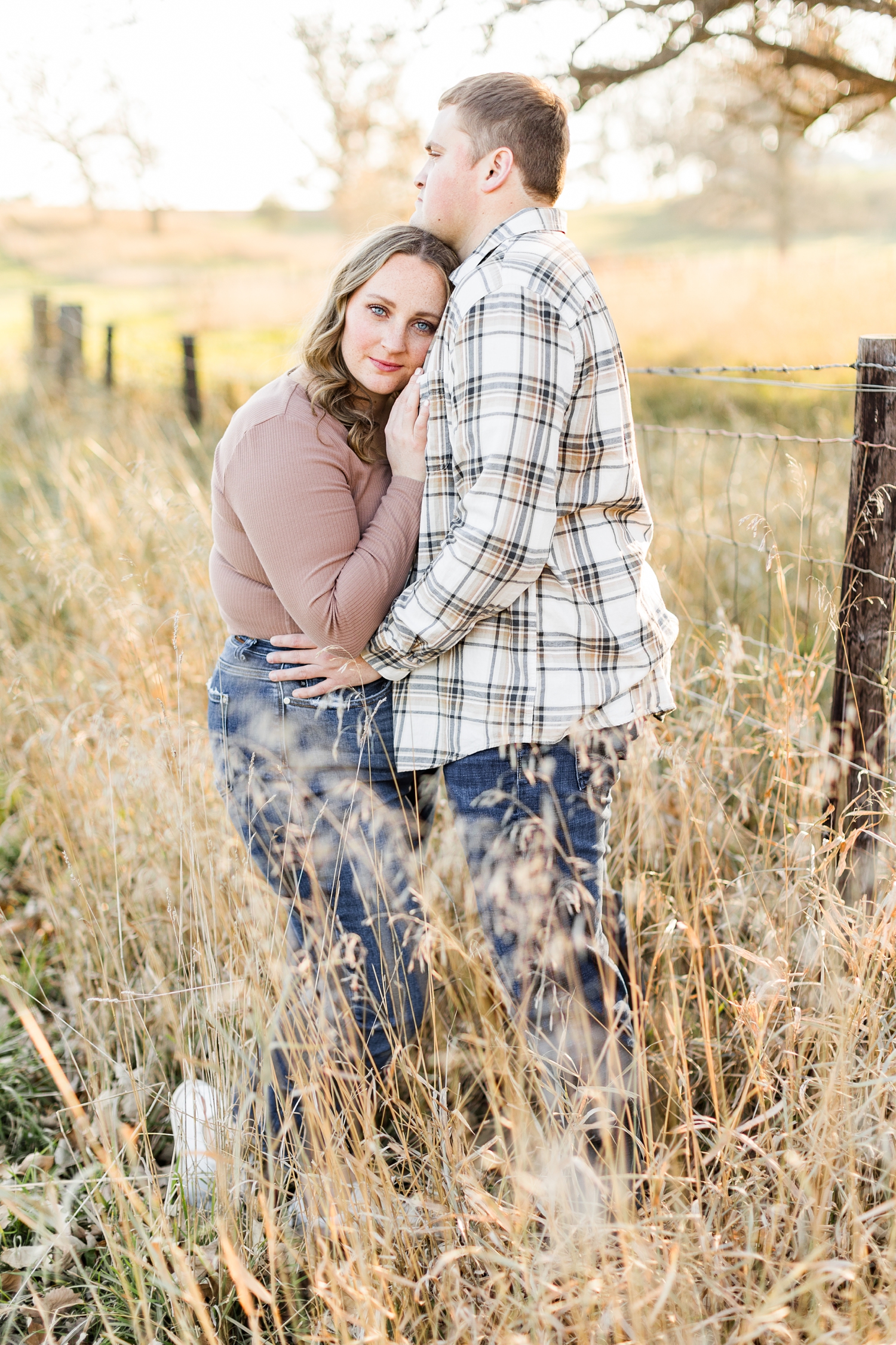 Shelby and Taylor embrace each other along a fence line in a grass pasture during golden hour in Iowa | CB Studio