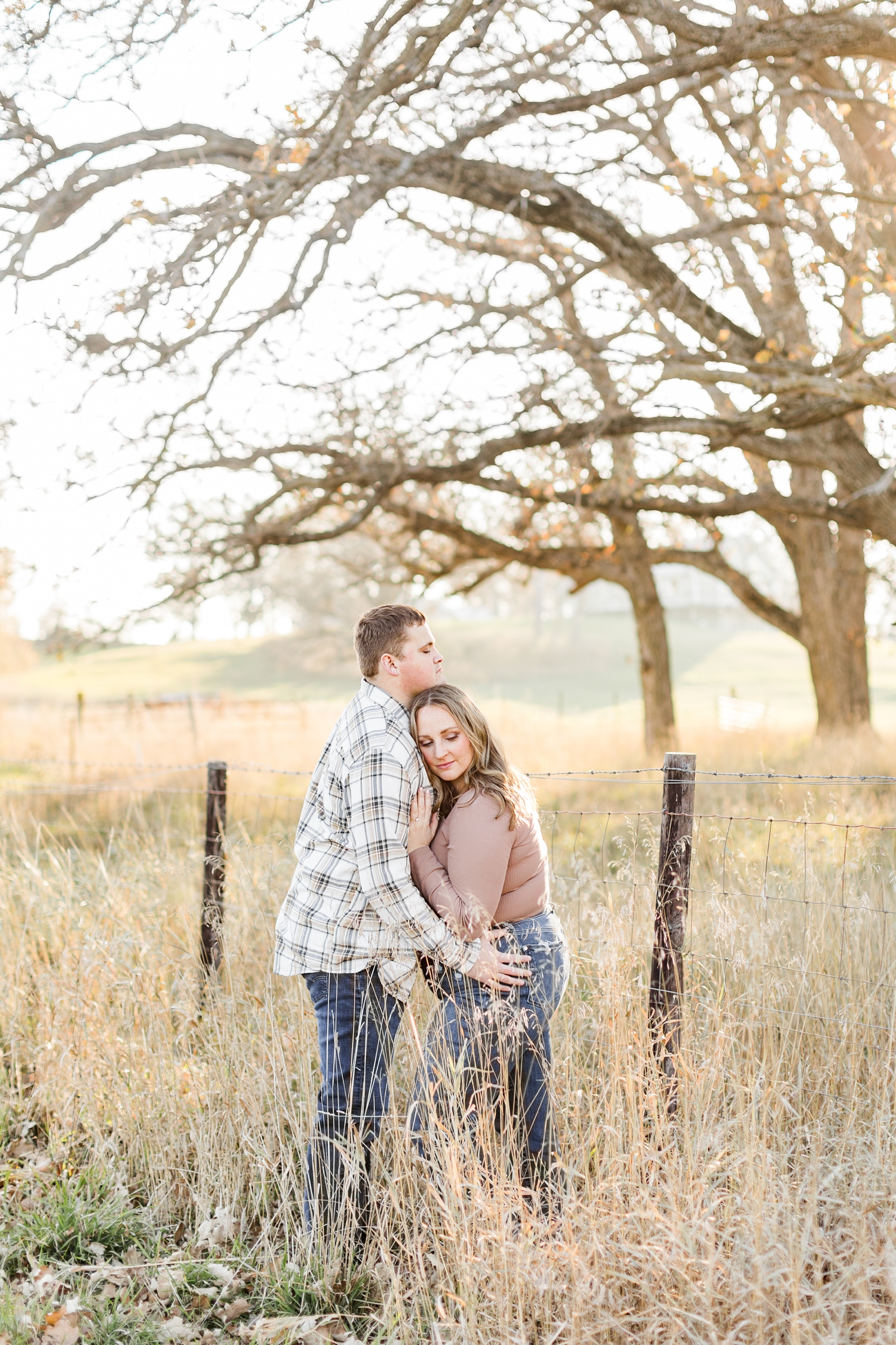 Shelby and Taylor embrace each other along a fence line in a grass pasture during golden hour in Iowa | CB Studio