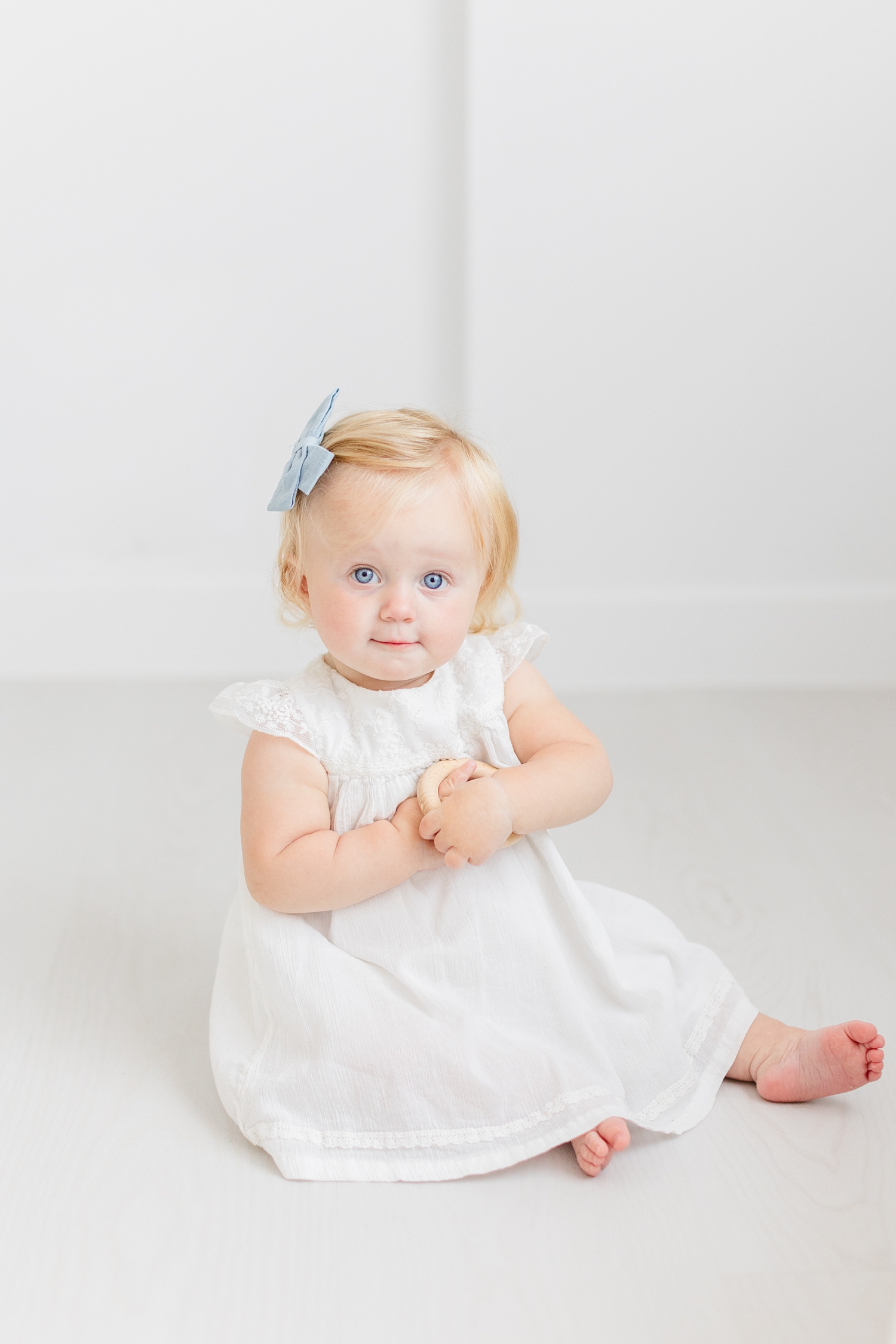 Baby Ryer dressed in an antique white dress plays with a wooden toy in an all white room | CB Studio