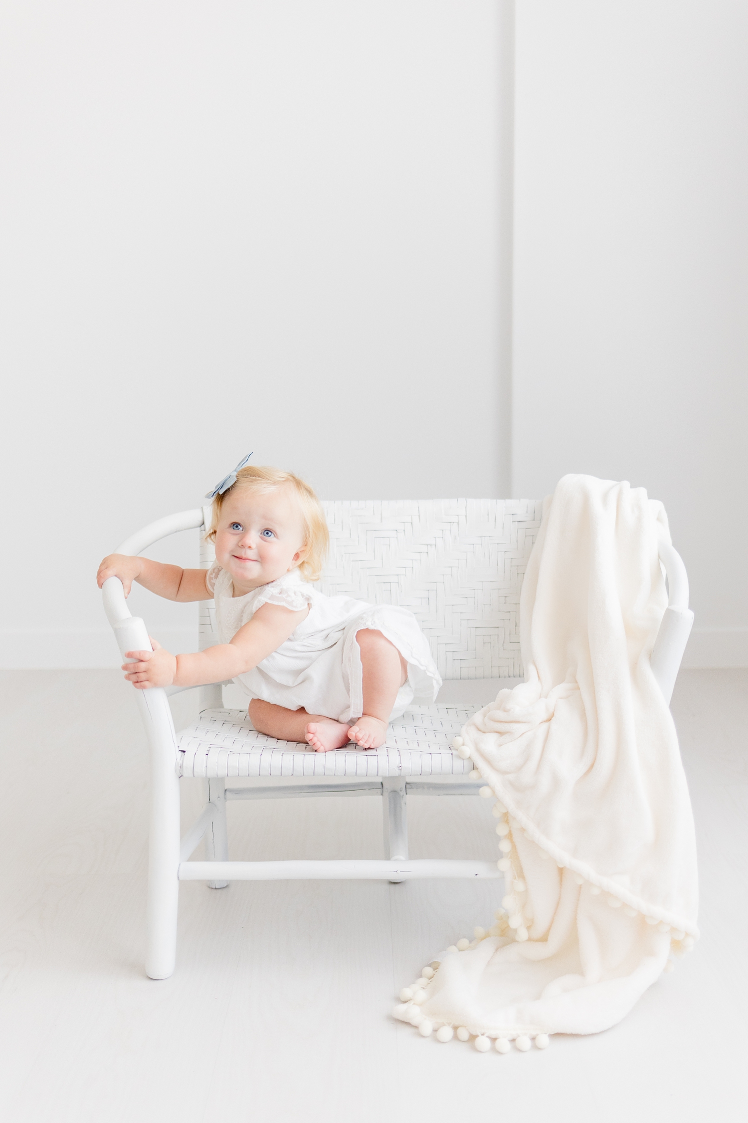 Baby Ryer dressed in white sits on a white wicker bench in a solid white room | CB Studio