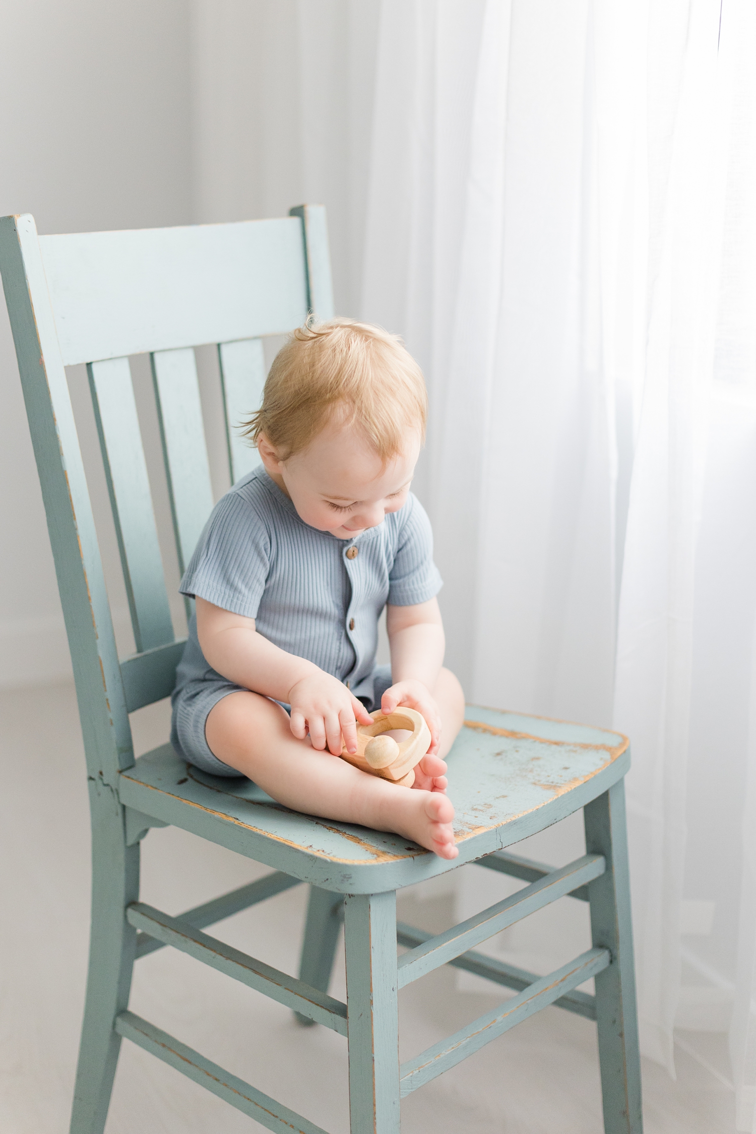Baby Lewis sits in a blue chair playing with a wooden toy in front of a window draped in white curtains | CB Studio