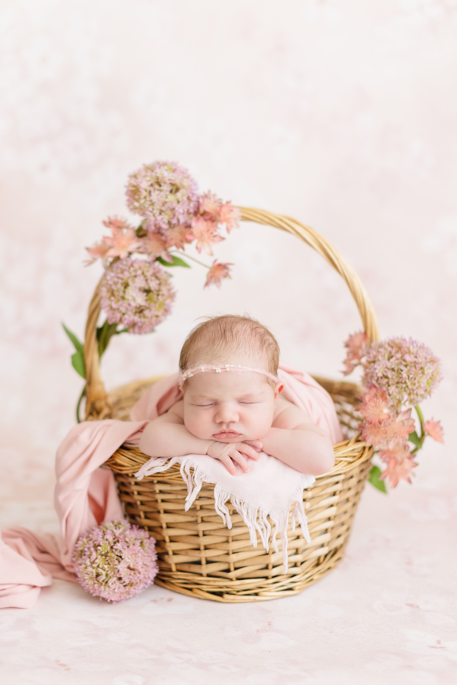 Baby Grace dressed in pink sleeps peacefully in a wicker basket surrounded by pink florals | CB Studio