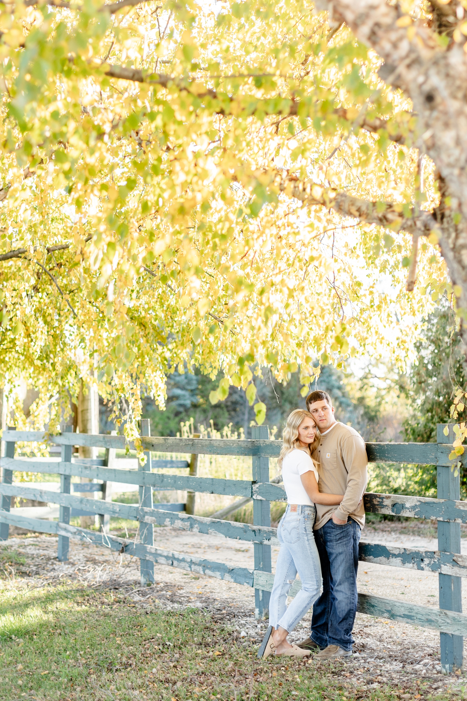Reed leans against a teal wooden fence as Nicole hugs him close, surrounded by yellow birch tress in the fall at River Valley Winery | CB Studio