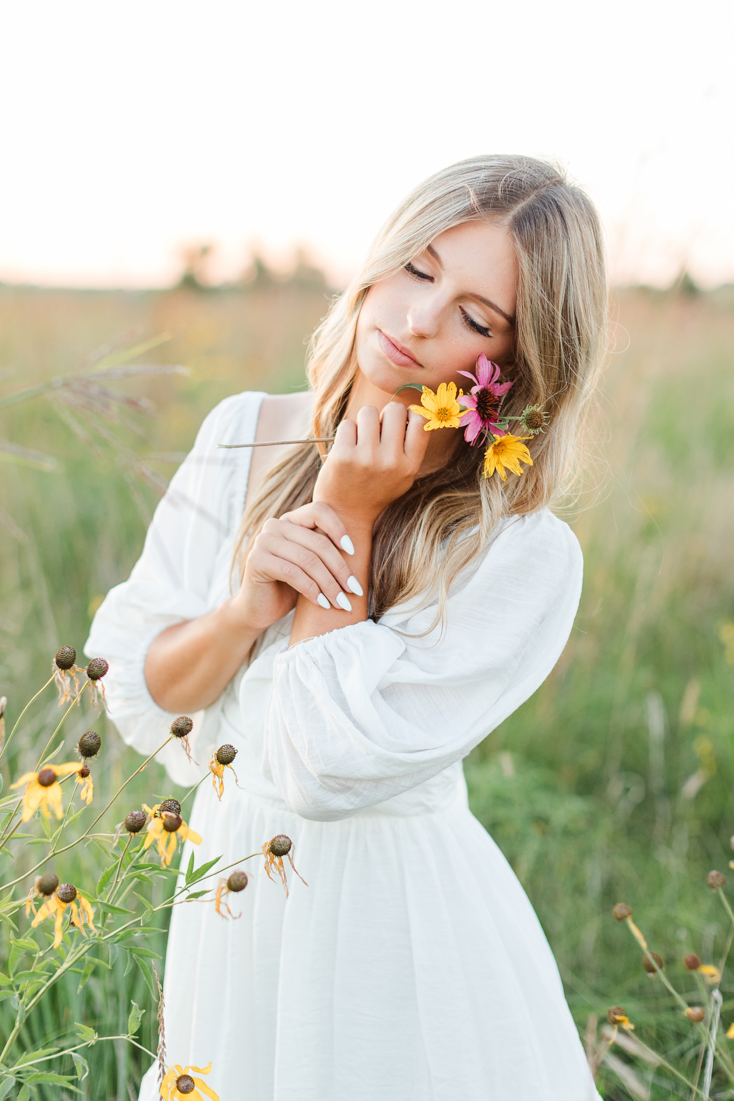 Eden, wearing a white dress, holds a small wildflower bouquet to her cheek as she stands in a flower field at sunset | CB Studio