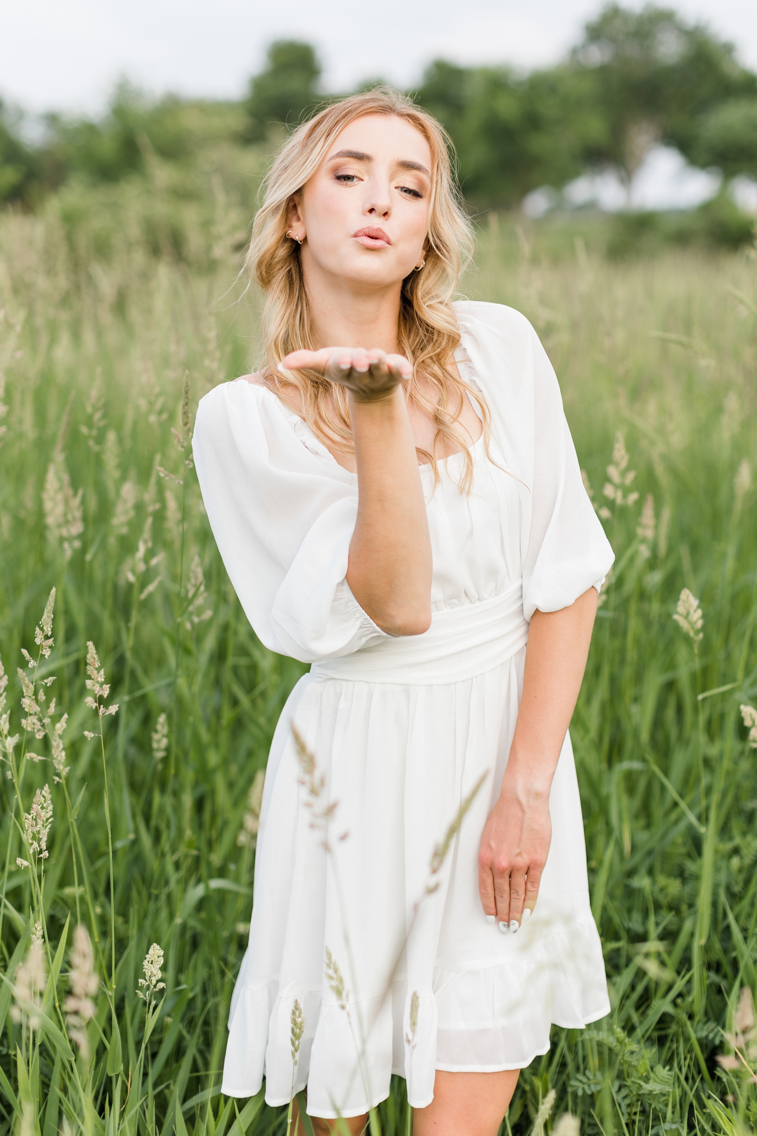 Raegan wearing a white dress blows a kiss as she stands in a tall grassy field | CB Studio