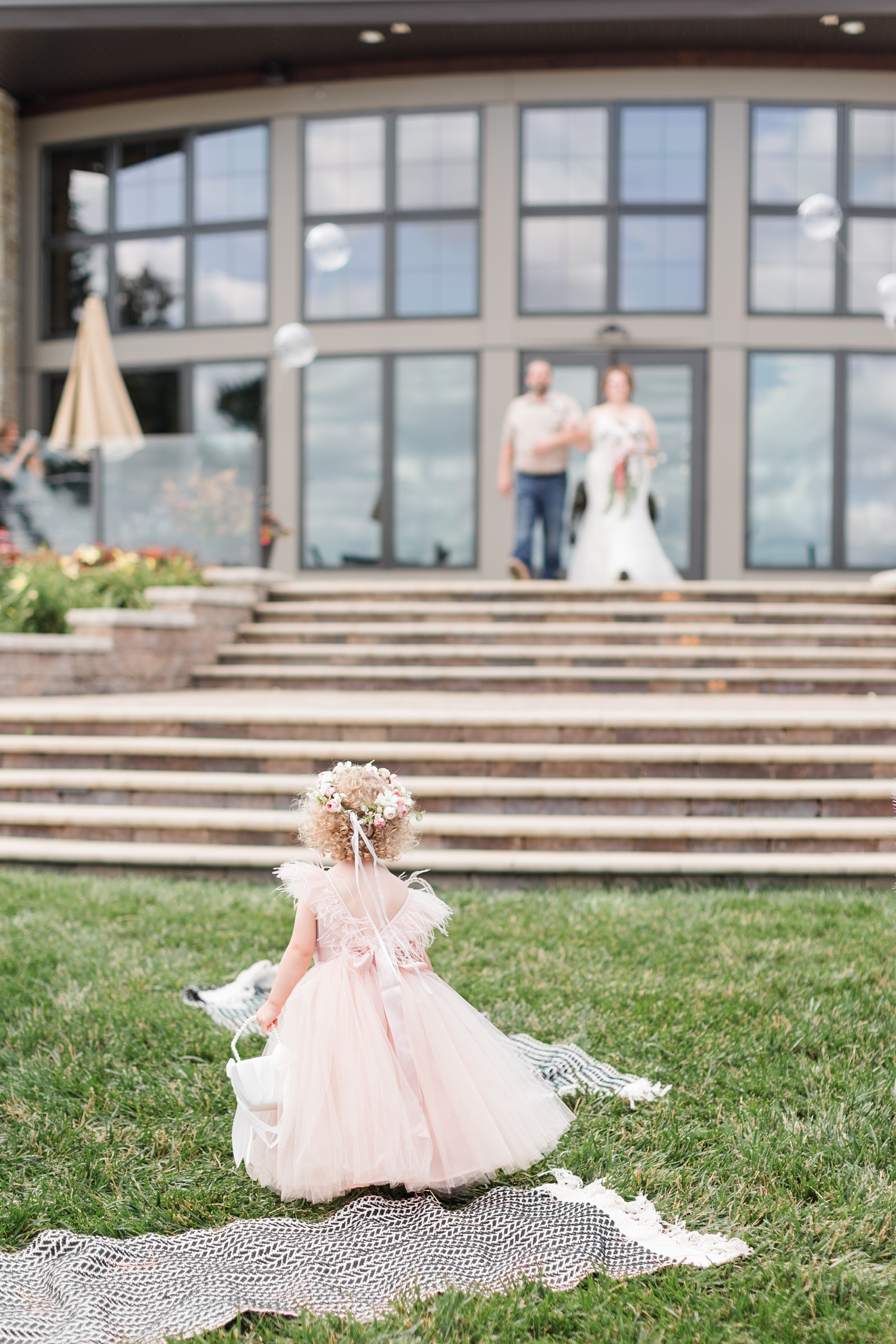A young flower girl dressed in blush and surrounded by bubbles, stands at the end of the aisle and watches as the bride walks down with her father in the background | CB Studio
