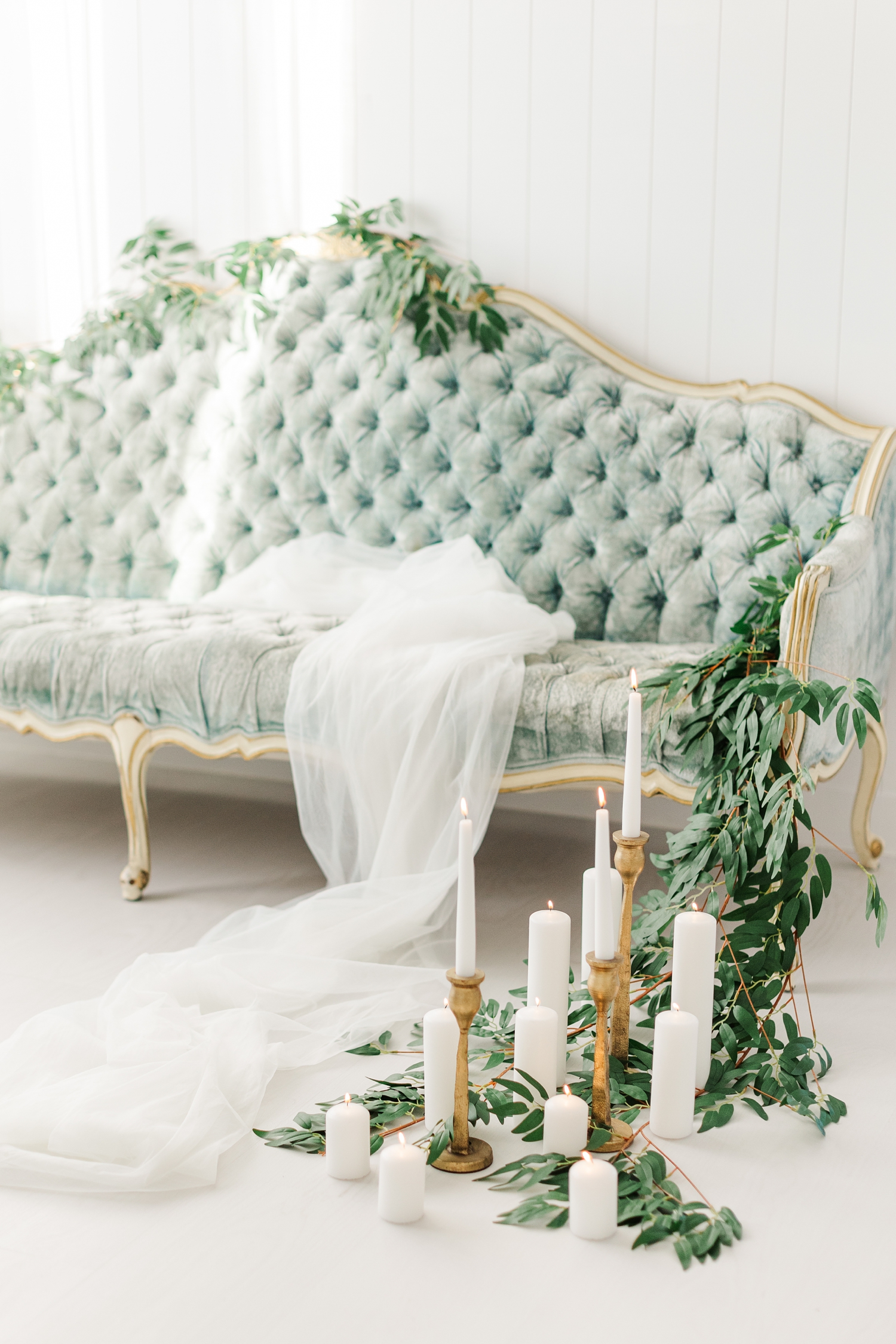 A cathedral length veil is draped across an antique, tufted, blue, victorian style couch draped in greenery and surrounded by white pillar candles and gold candlesticks | CB Studio
