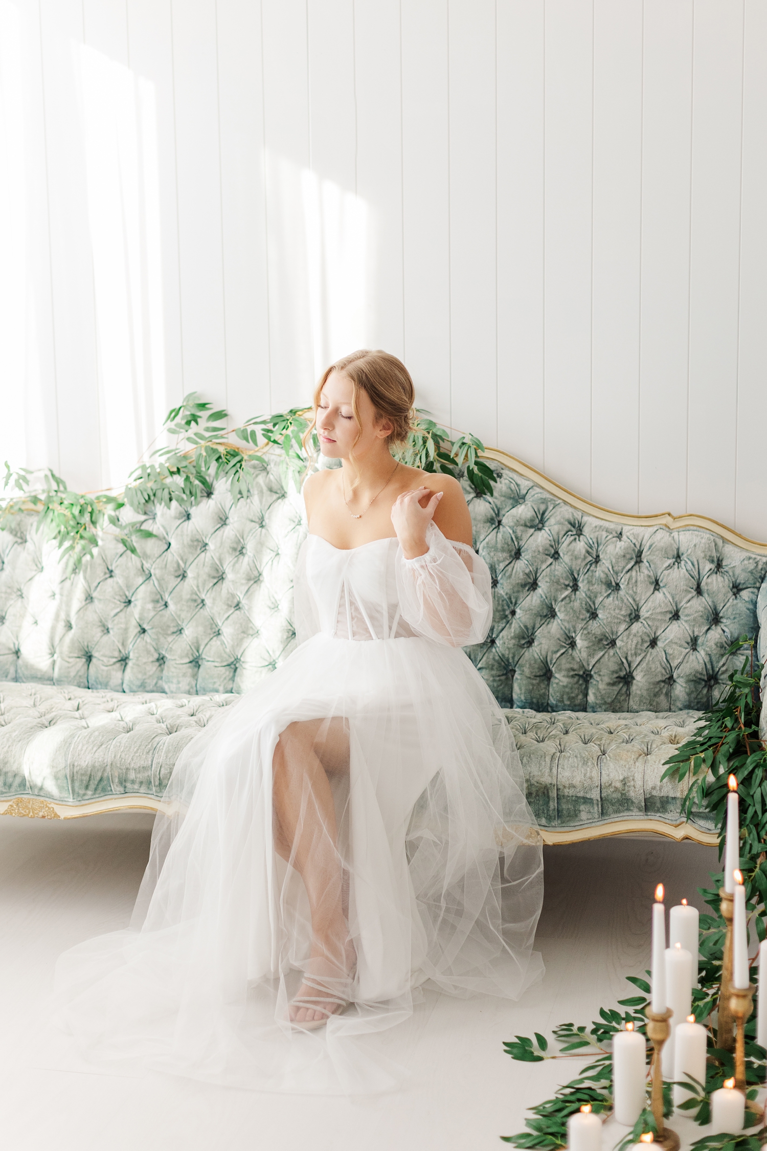 Bride, Ryley dressed in her vintage style wedding gown, gracefully sits on an antique, tufted, blue, victorian style couch draped in greenery | CB Studio