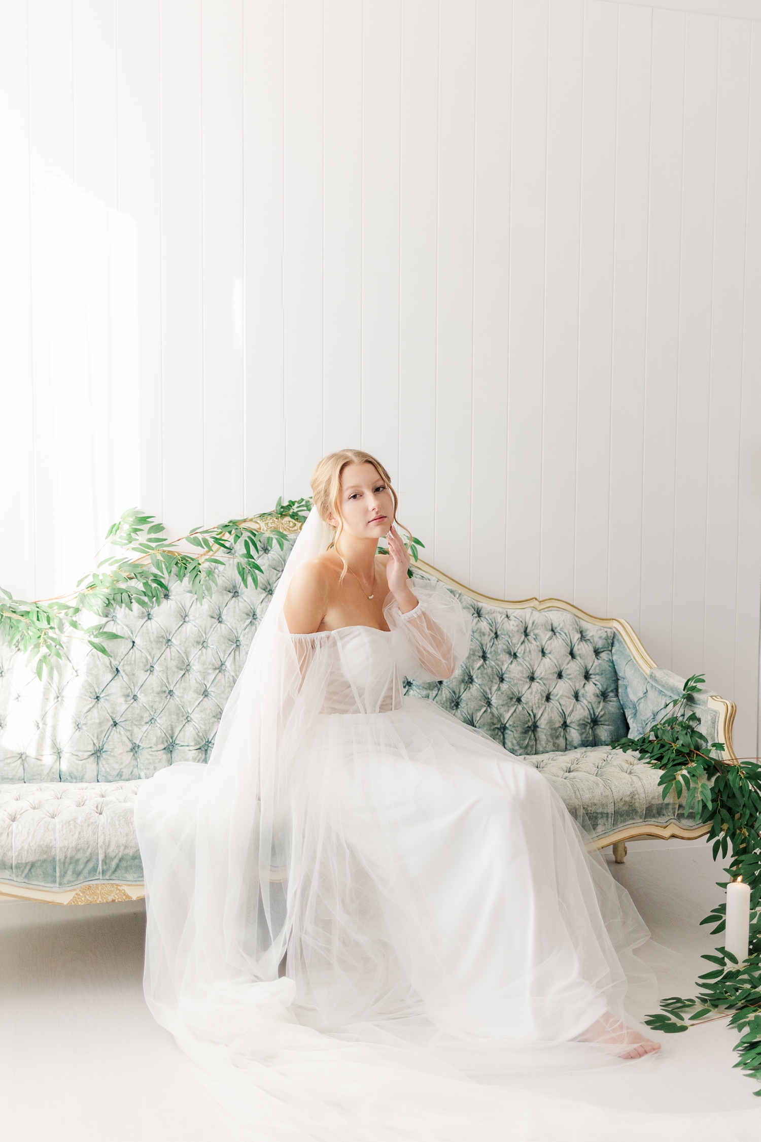 Bride, Ryley dressed in her vintage style wedding gown, gracefully sits on an antique, tufted, blue, victorian style couch draped in greenery | CB Studio