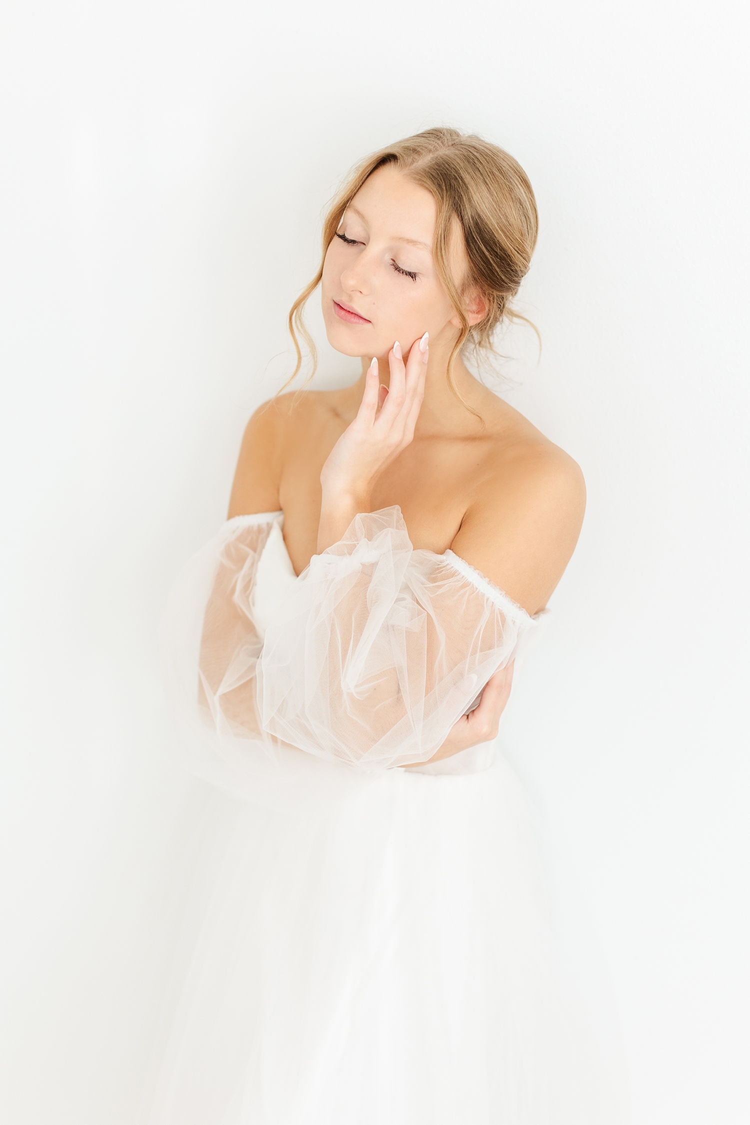 Bride, Ryley, leans against a white wall as she gracefully touches her cheek | CB Studio