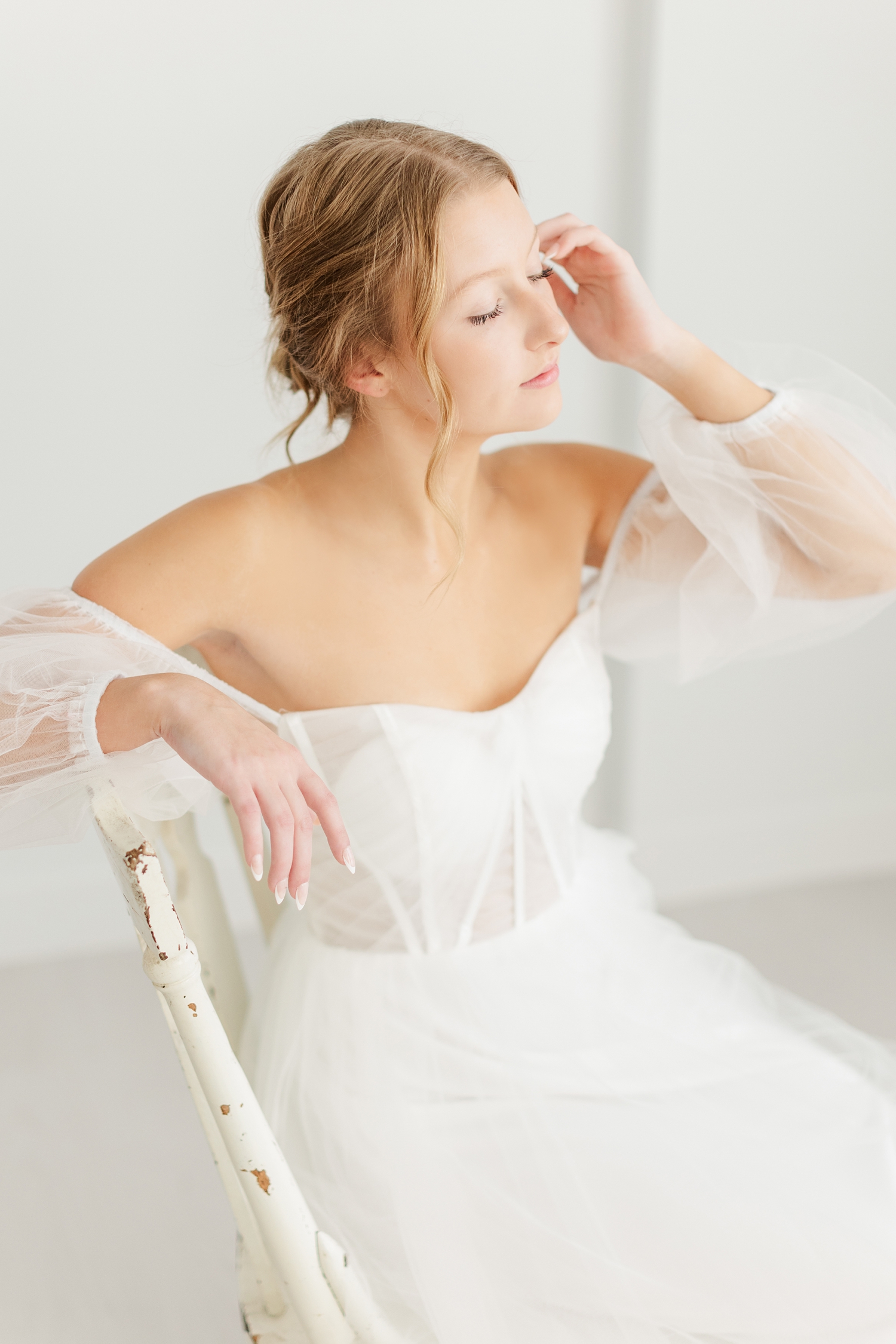 Bride, Ryley, gracefully sits in an antique wooden chair as she gently tucks her hair back | CB Studio