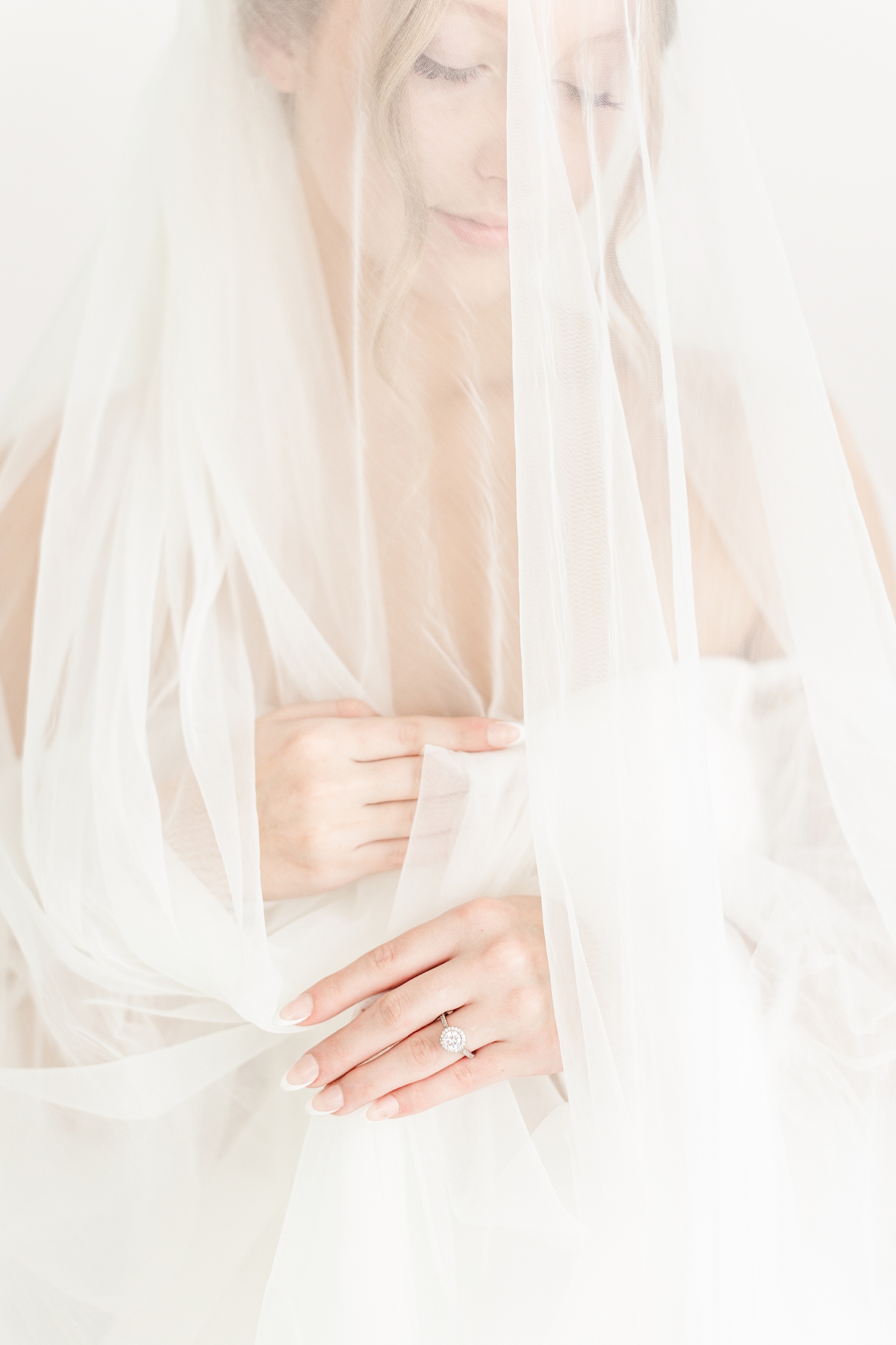 Bride, Ryley, draped in her cathedral length veil, holds the veil up to her chest as she looks down at her engagement ring | CB Studio
