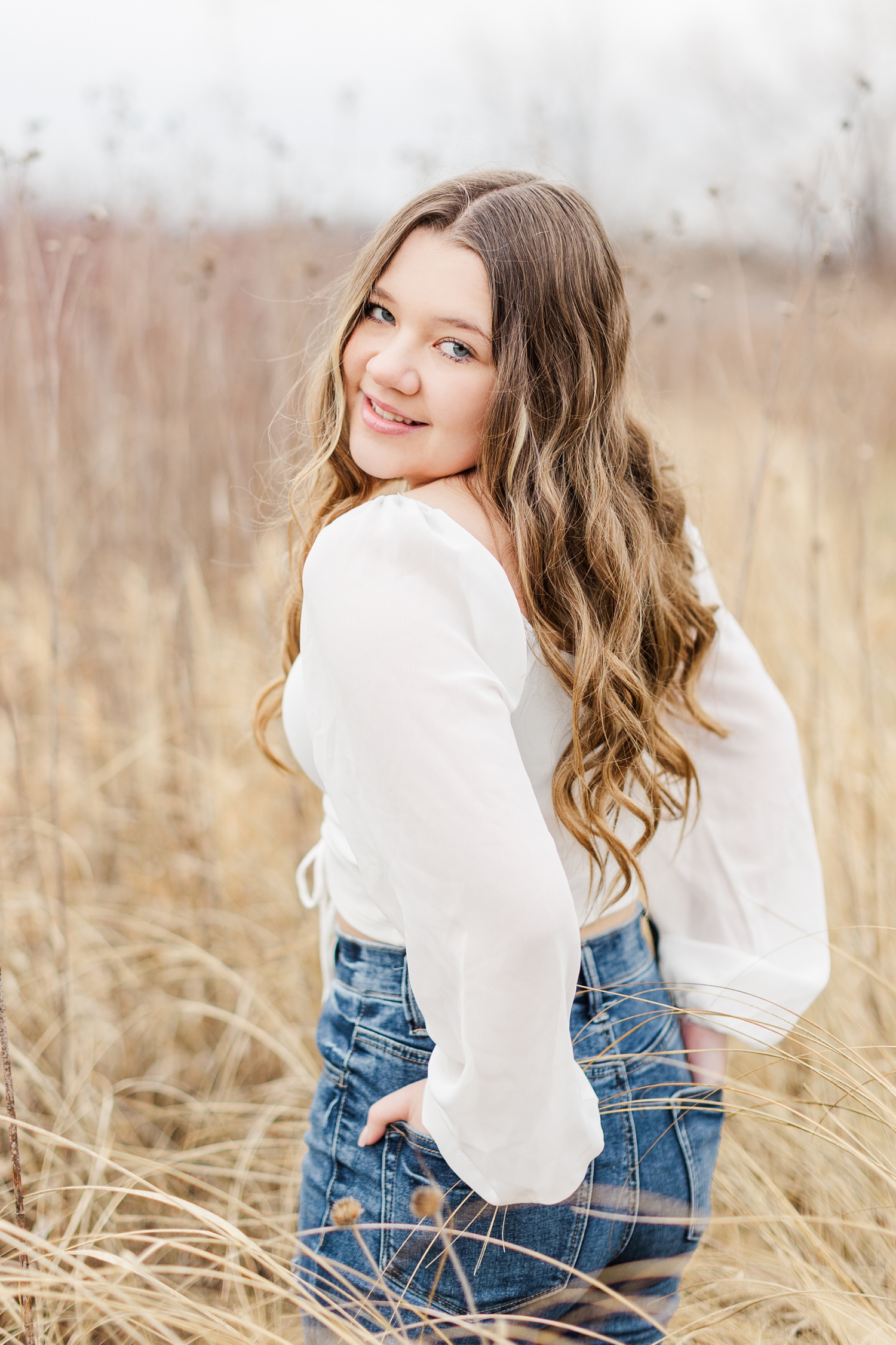 Sierra looks back over her shoulder with her hands in her back jean pockets while standing in a dead grassy field at Water's Edge Nature Center | CB Studio