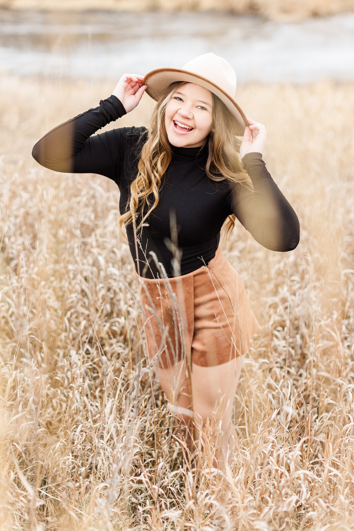 Sierra laughs as she stands in a dead grassy field, wearing a camel colored skort with a black turtleneck bodysuit and a camel colored, wide brim hat from West Vendee | CB Studio