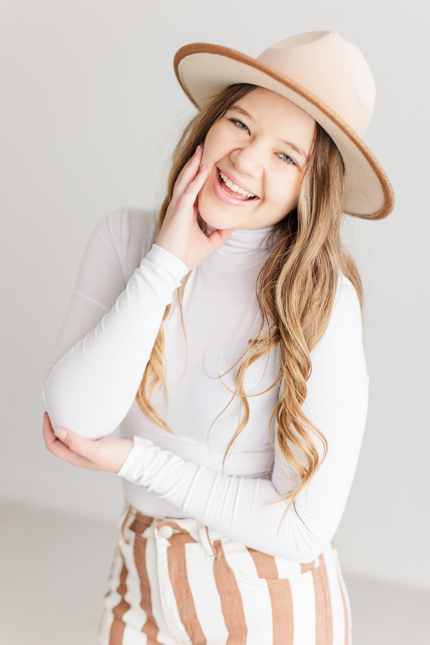 Sierra laughs as she tilts her head, wearing white and camel striped bell bottoms with a white turtleneck bodysuit and a camel colored, wide brim hat from West Vendee | CB Studio