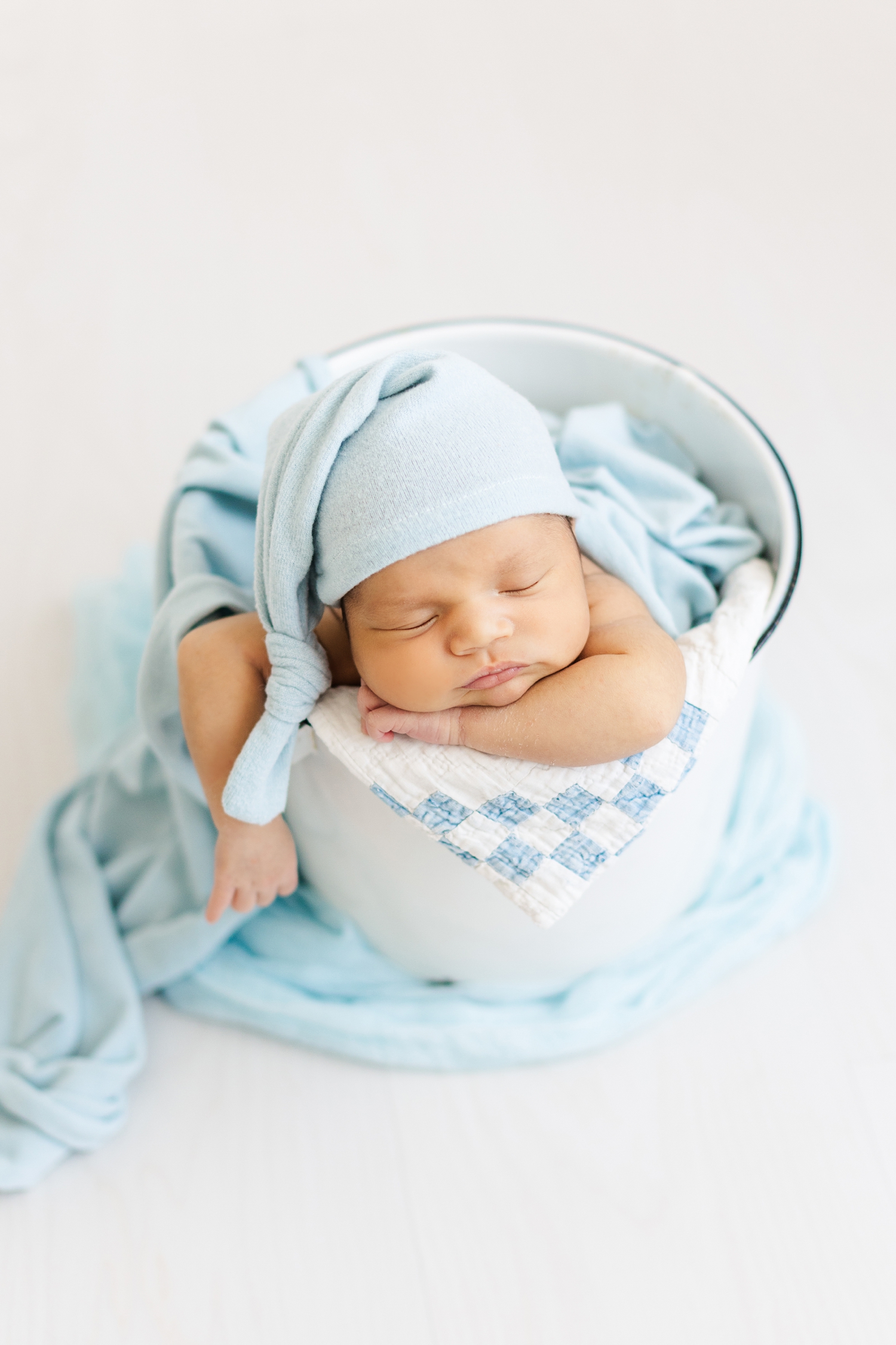 Baby Zachary nestled in a white bucket with a white and blue patch work quilt wearing a blue night cap and a blue swaddle draped over him and the bucket | CB Studio