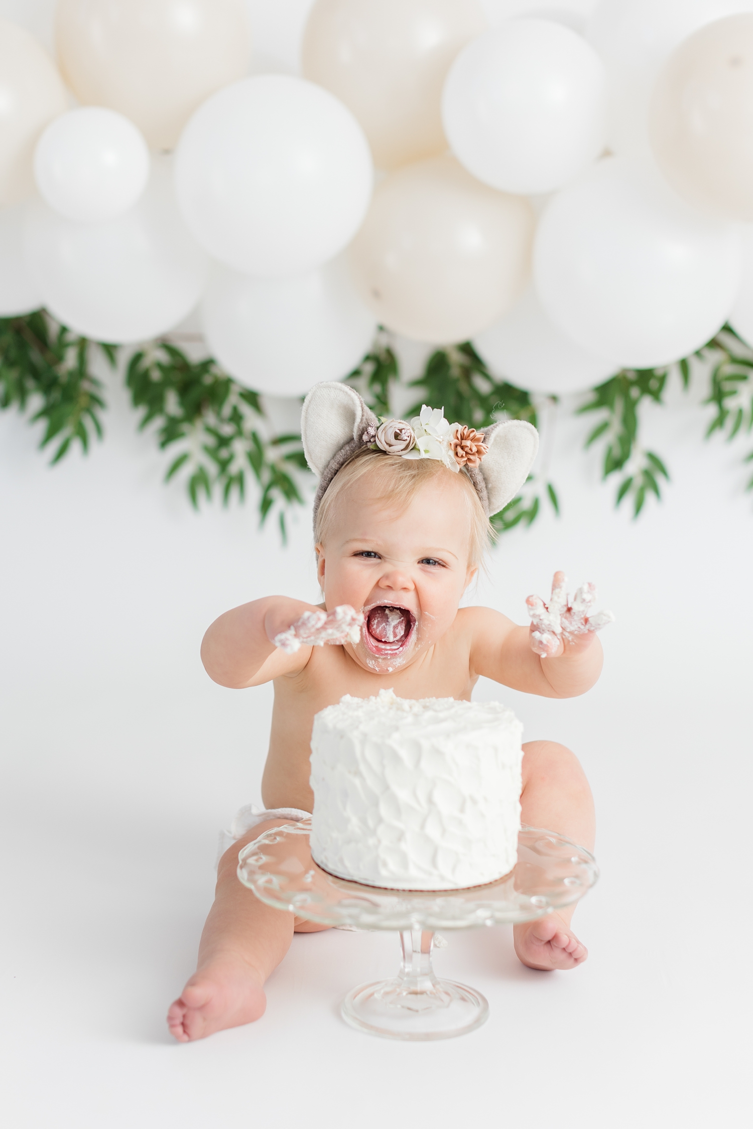 Baby Emersyn enjoys a white cake with a white balloon garland background to celebrate her first birthday | CB Studio