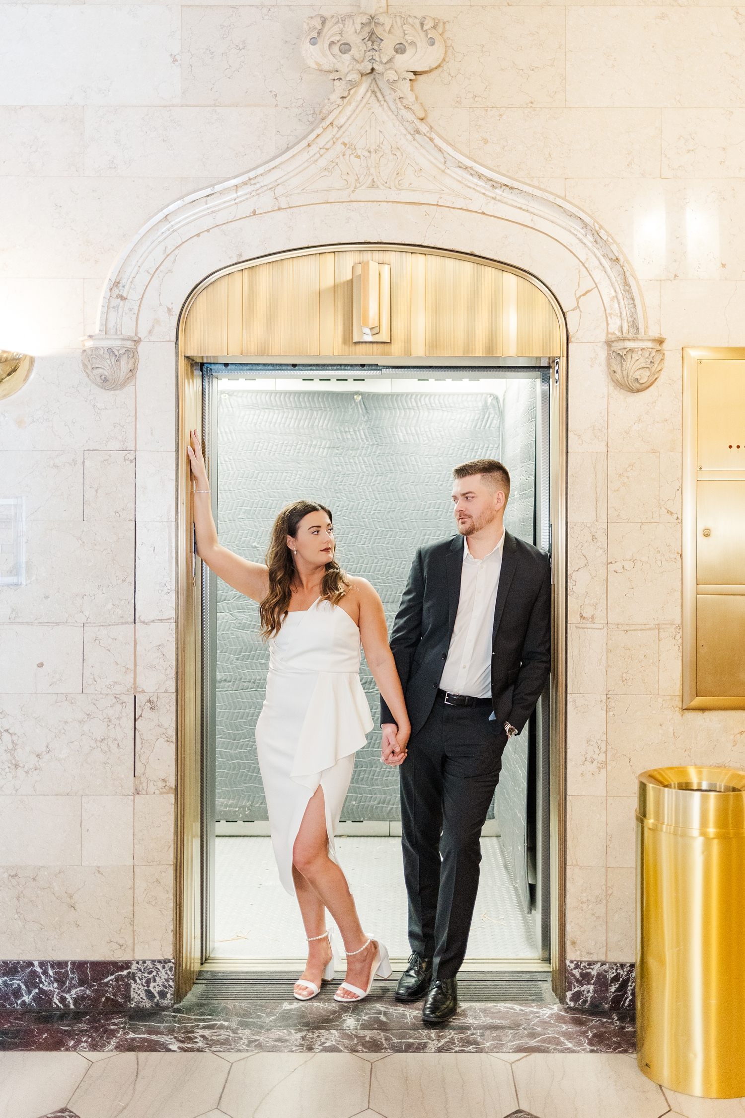 Jenna and Dustin lean against opposite frames of a gold elevator with the door open in the lobby entrance of The Equitable Building in Des Moines, IA | CB Studio