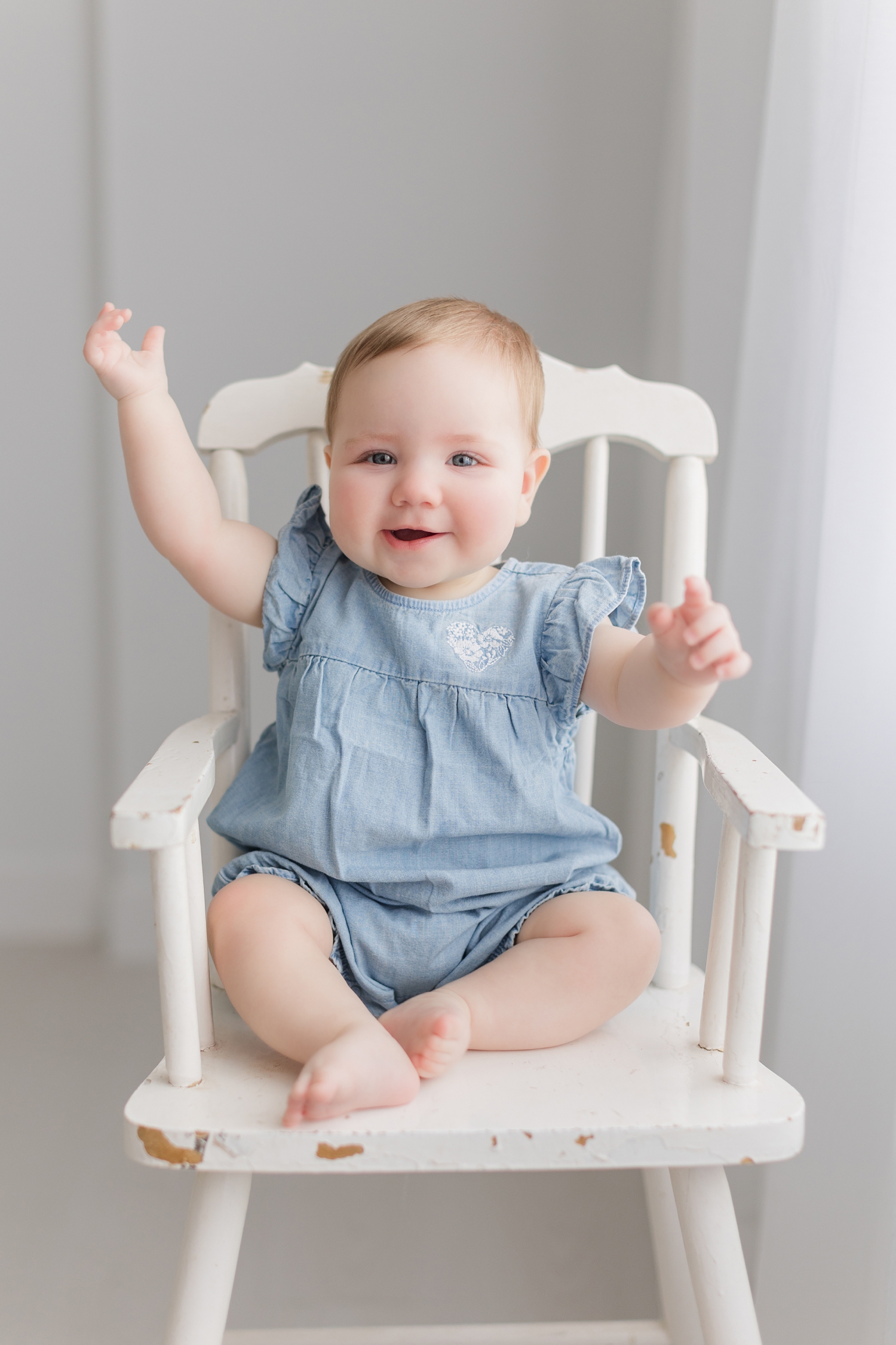 Baby Grace reaches her arms up and smiles as she sits on a vintage white high chair in an all white room. She is wearing a light blue chambray romper that matches perfectly with her blue eyes | CB Studio