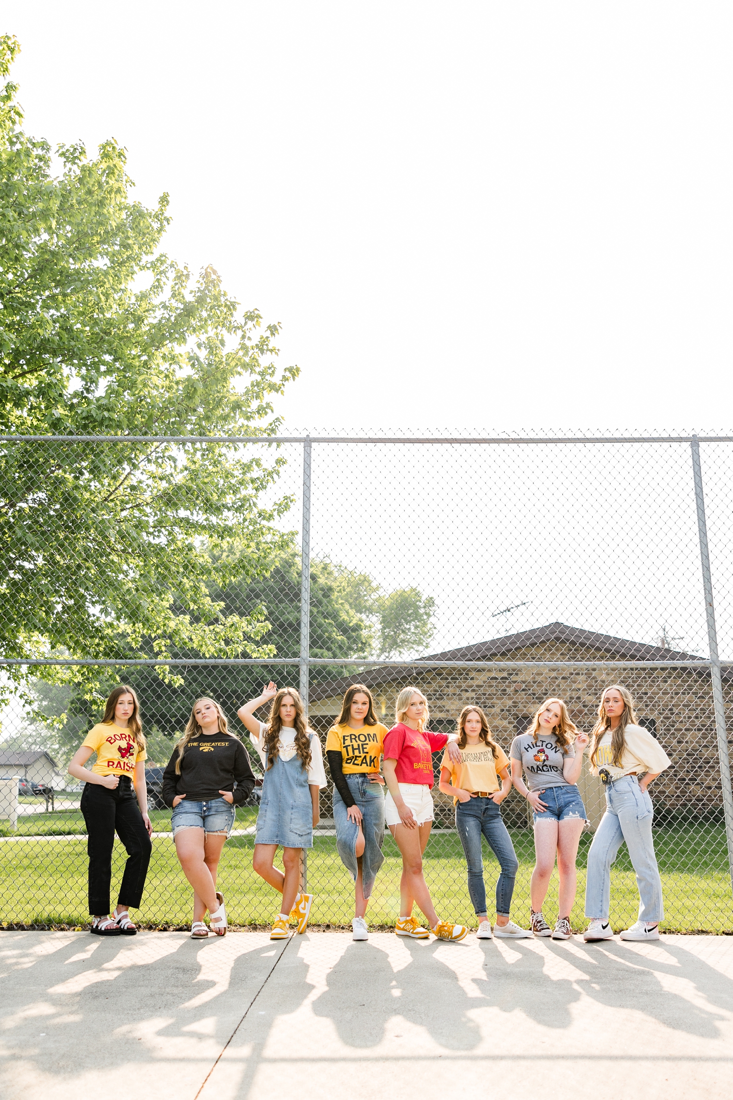 TEAM 25, all wearing Iowa or Iowa State clothing, model pose in a straight line as they lean against a chainlink fence | CB Studio