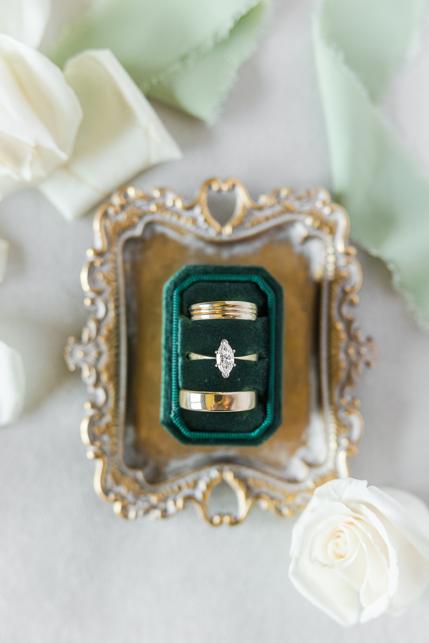 Gold wedding bands and a marquee cut solitaire engagement ring sit nestled in an emerald green Mrs. Box sitting on top of a gold ornate tray surrounded by white roses and sage green ribbon | CB Studio