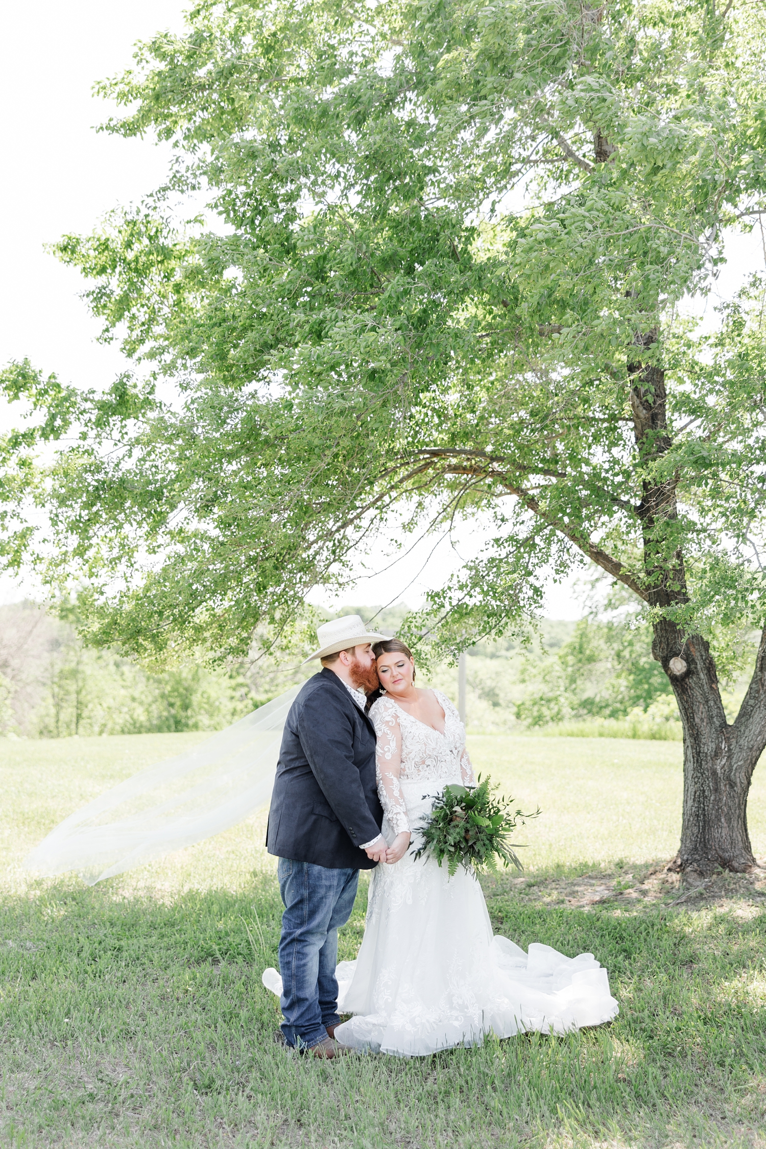 Trent kiss his new bride, Neely, on the cheek under a beautiful tree at Lizard Creek Ranch in Fort Dodge, IA | CB Studio