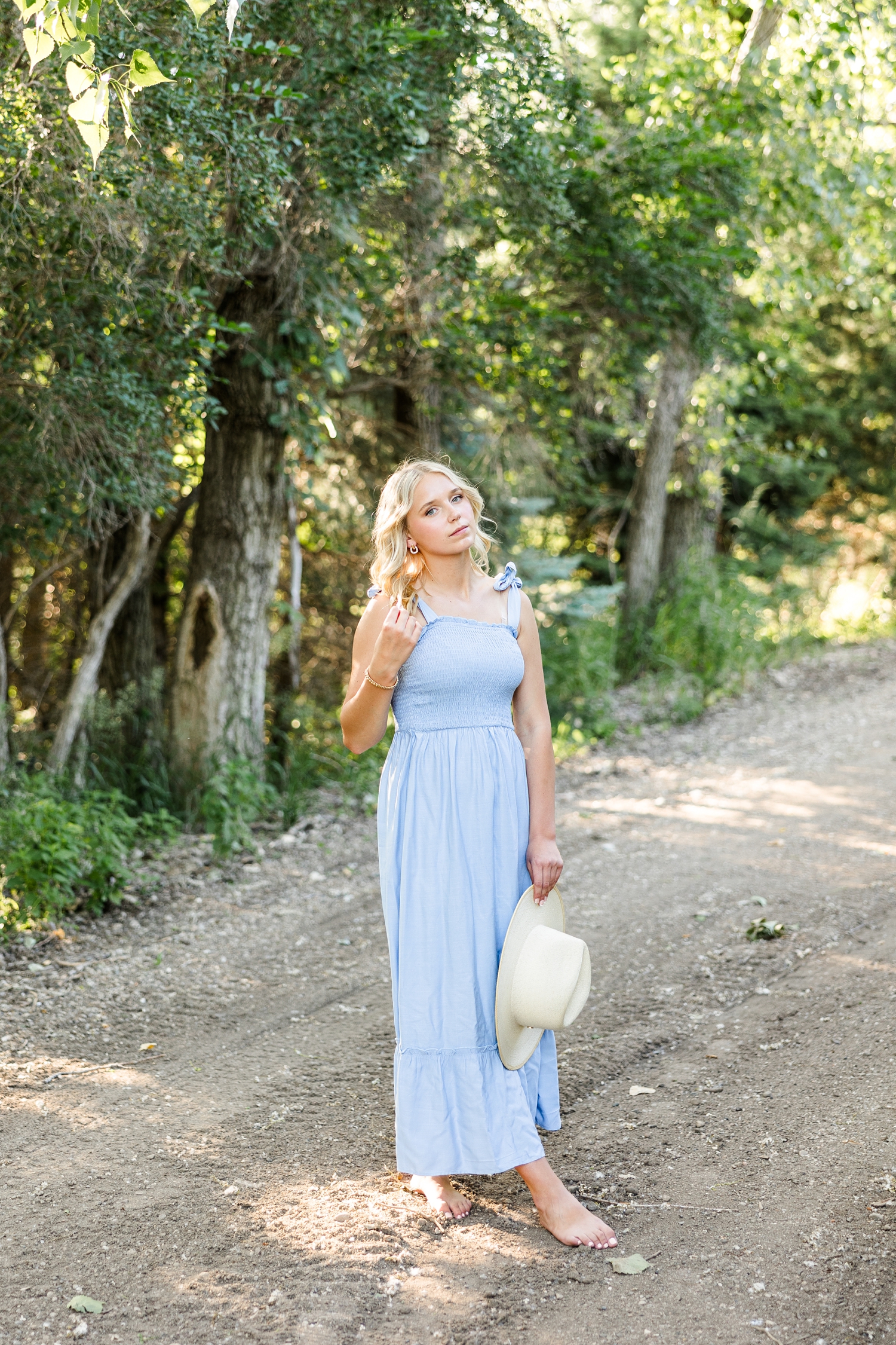 Addi, wearing a long blue dress and straw hat, looks to the camera with a blank stare as she stand along a path at Wild Haven | CB Studio