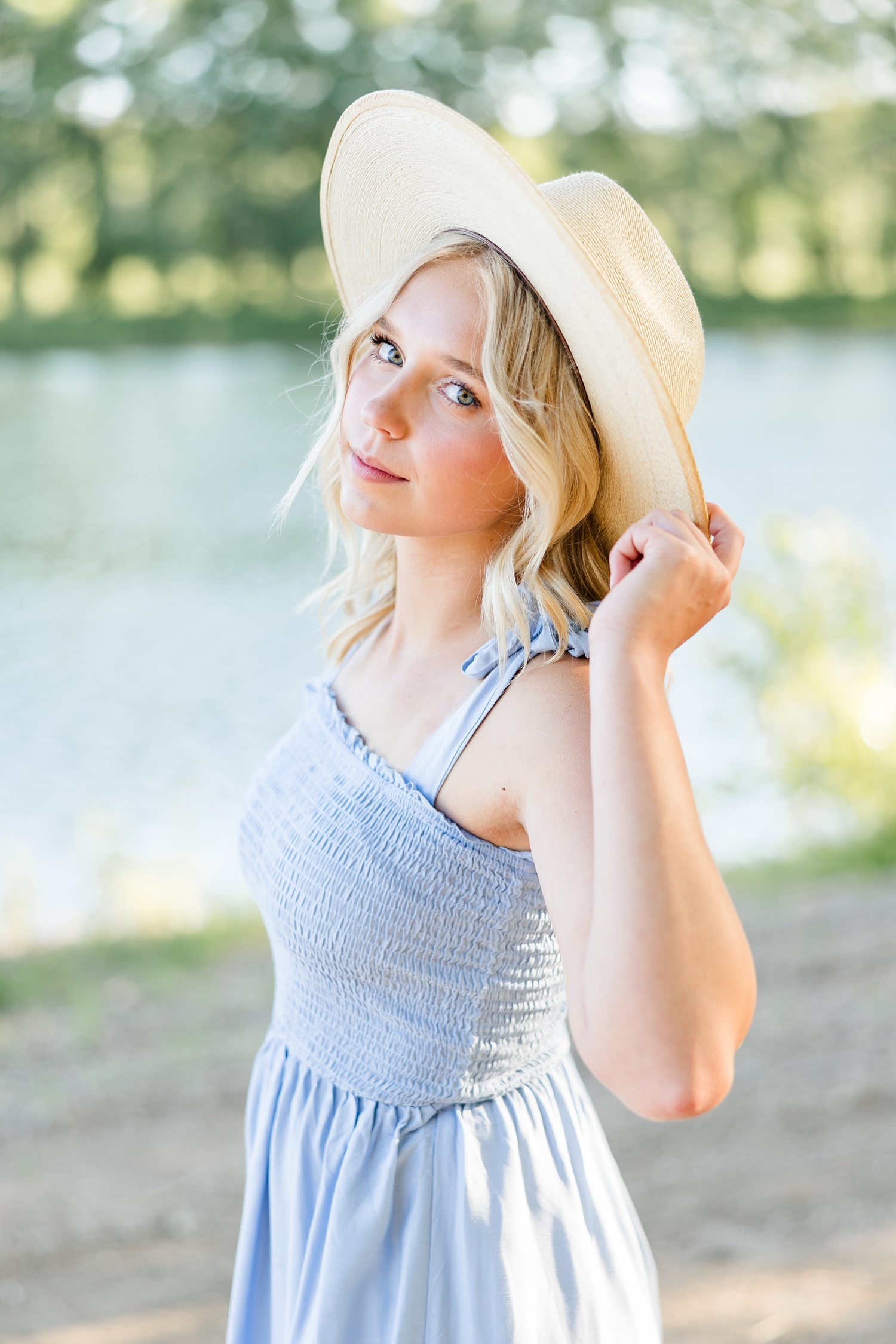 Addi, wearing a long blue dress and straw hat, looks over her shoulder while she pinches the hat at Wild Haven | CB Studio