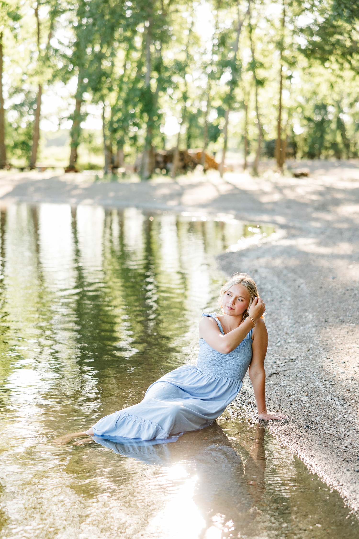 Addi, wearing a long blue dress, sits in the water while she brushes her hair aside on the edge of a pond at Wild Haven | CB Studio