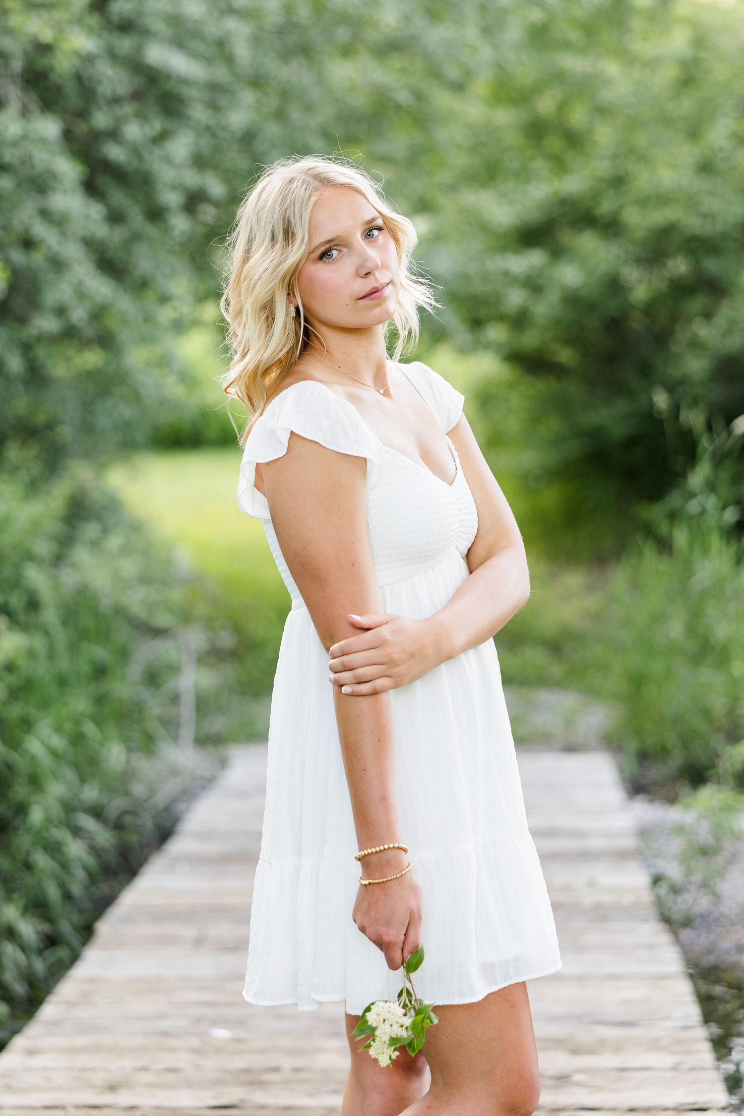 Addi, wearing a white dress, stands on a wooden bridge at Water's Edge Nature Center while holding a white flower | CB Studio