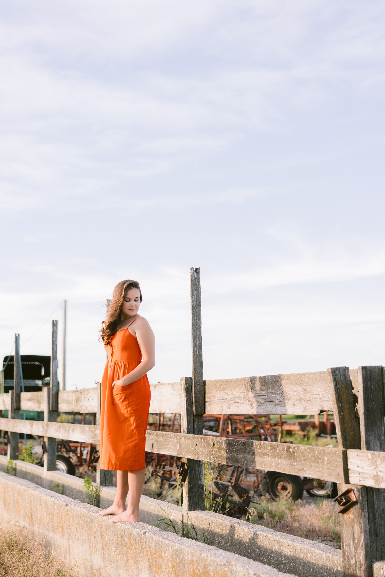 Adeline, wearing a reddish orange dress, stands on old concrete cattle bunks as she looks down over her shoulder | CB Studio