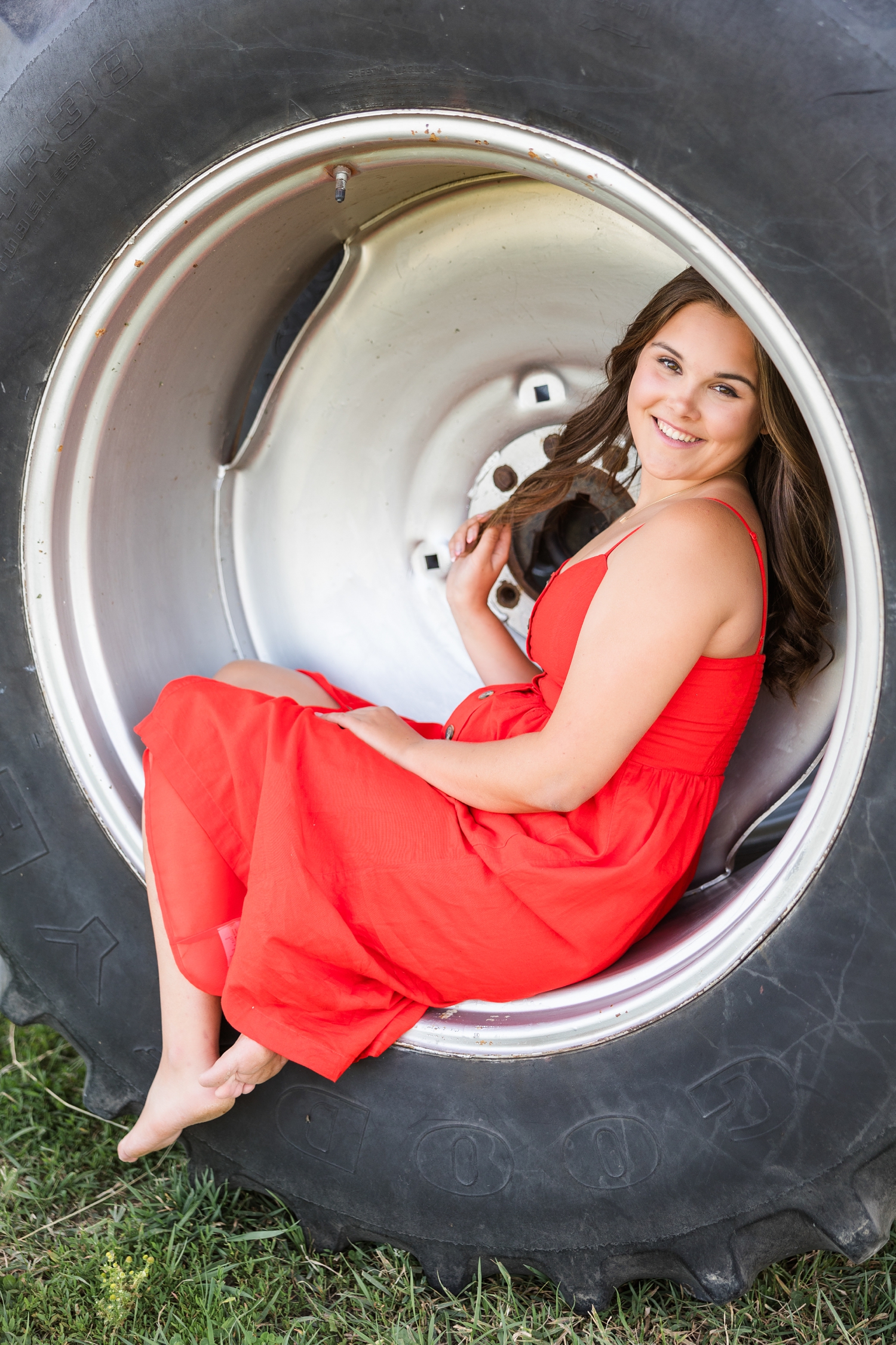 Adeline, wearing a red dress, sits in the hub of a tractor wheel while she holds a strand of her hair | CB Studio