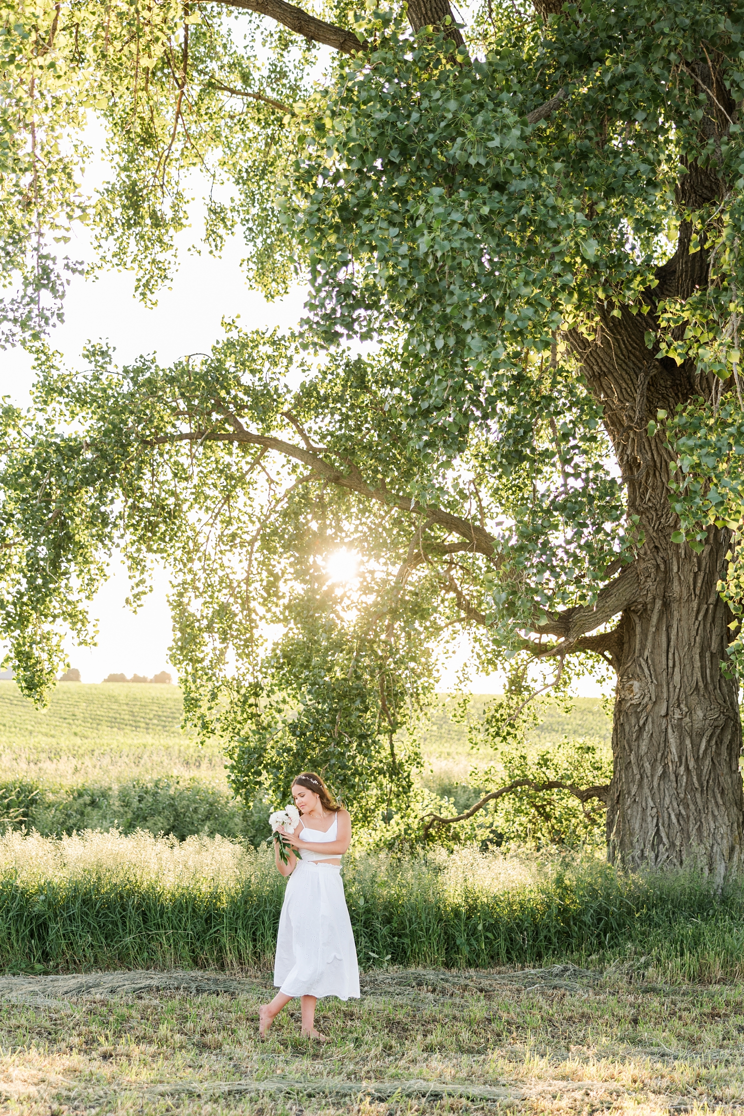 Adeline, wearing a white two piece skirt set, stands in a grassy field next to a giant oak tree at sunset holding a bouquet of white peonies as she softly looks down | CB Studio