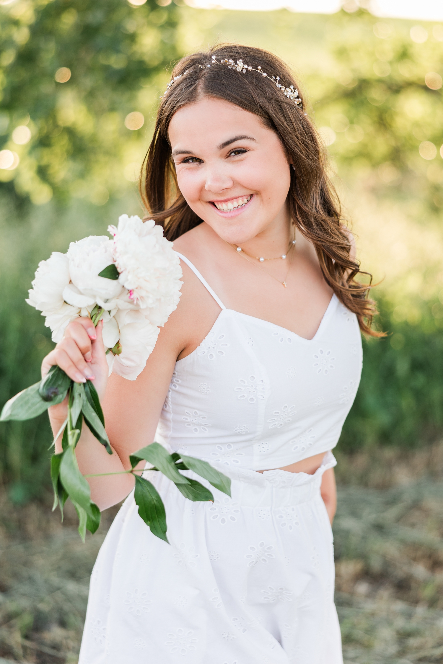 Adeline, wearing a white two piece skirt set, stands in a grassy field at sunset holding a bouquet of white peonies as she smiles | CB Studio