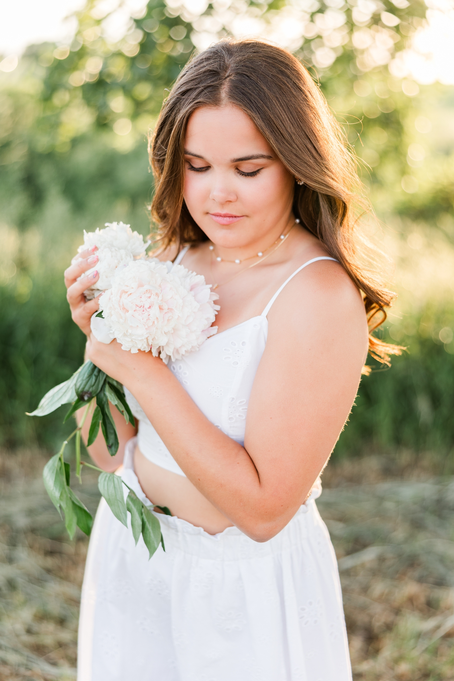 Adeline, wearing a white two piece skirt set, stands in a grassy field at sunset holding a bouquet of white peonies as she softly looks down | CB Studio